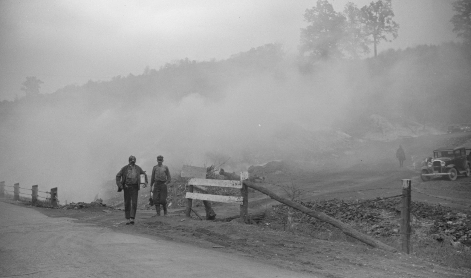 Black-and-white photograph of two miners walking with smoke behind them and another miner in the background.