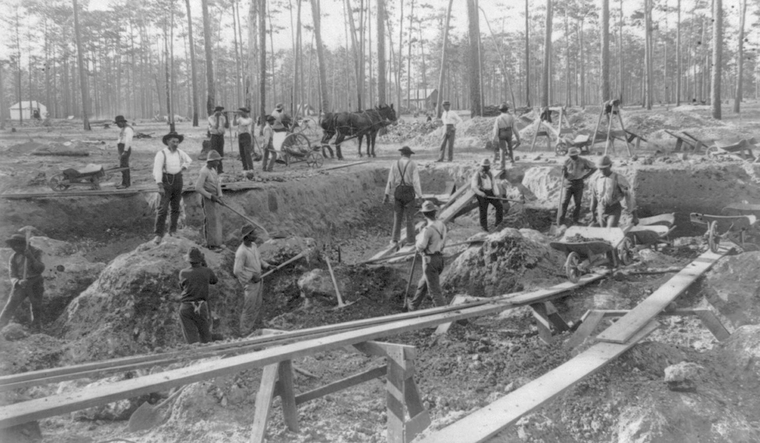 Black and white photograph of men working in a shallow phosphate mine, ca. 1890. The men stand in a shallow pit, many with picks and shovels.