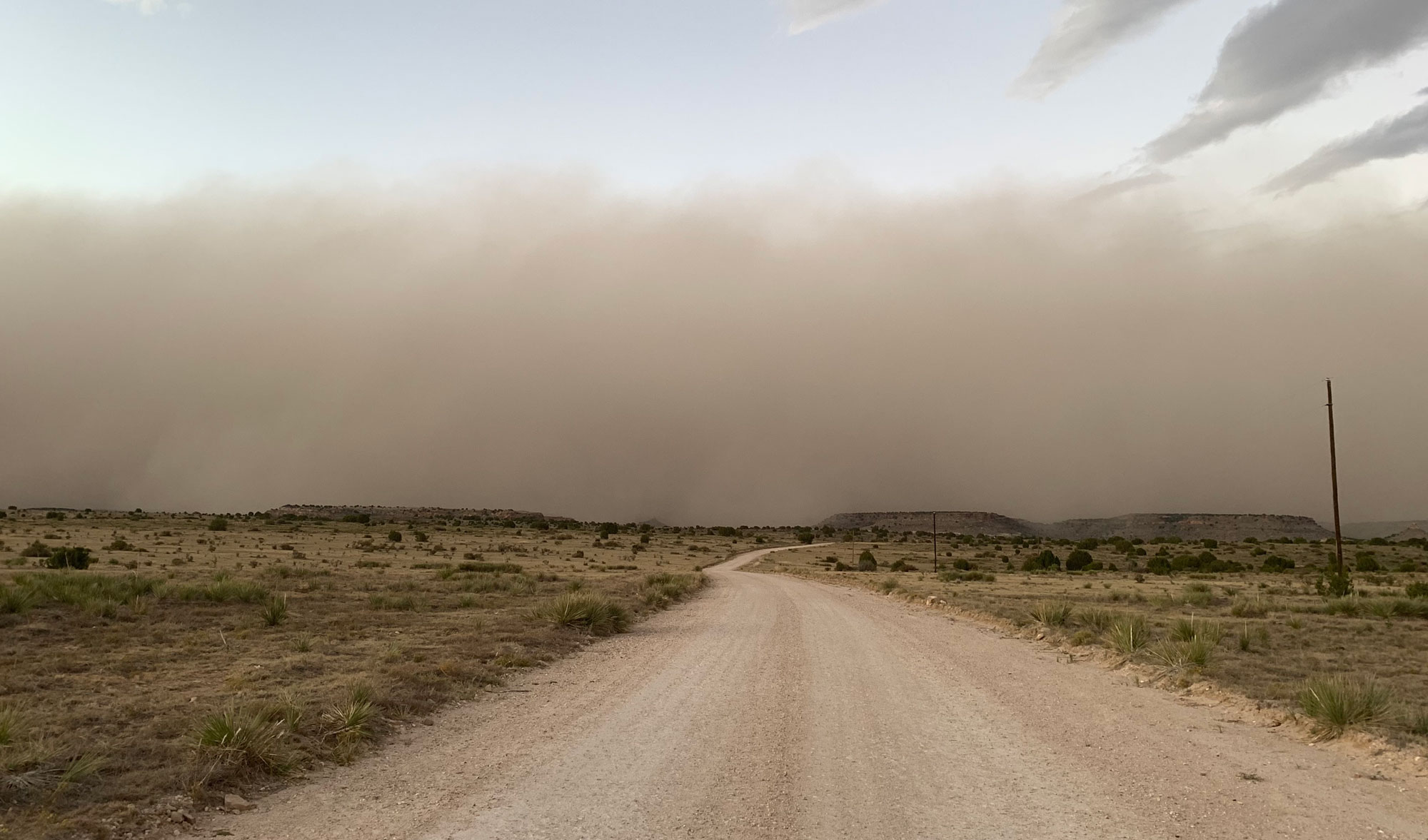 Photograph of a dust storm in Cimmaron County, Oklahoma, in 2011. The image shows a dirt road bisecting the foreground and veering right, with grass and yuccas on either side. On the horizon, a dust cloud can be seen over low hills.