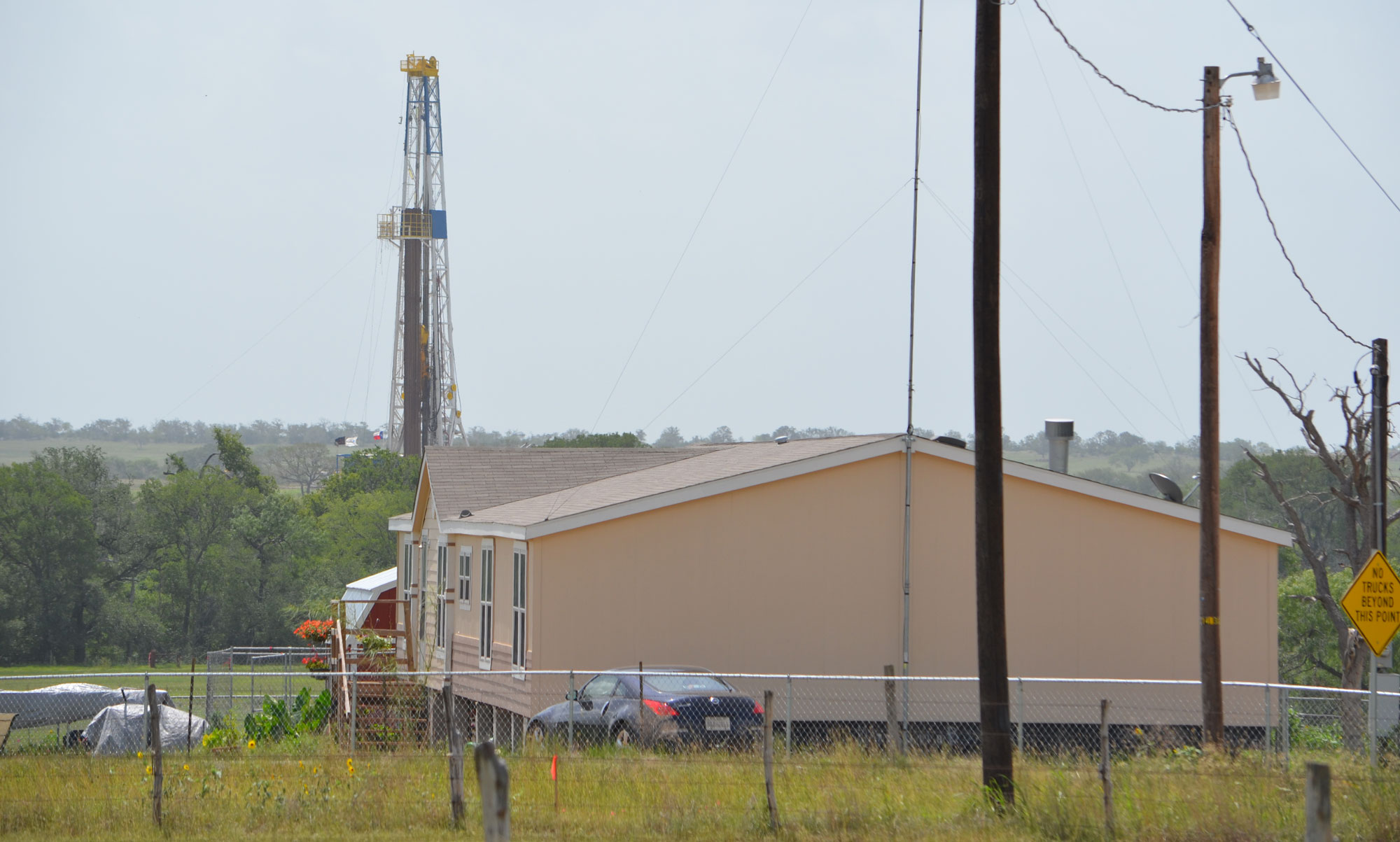 Photograph of a drill rig near a home in the Eagle Ford Shale. The home is in the foreground, the tower of the drill rig behind some trees in the background.