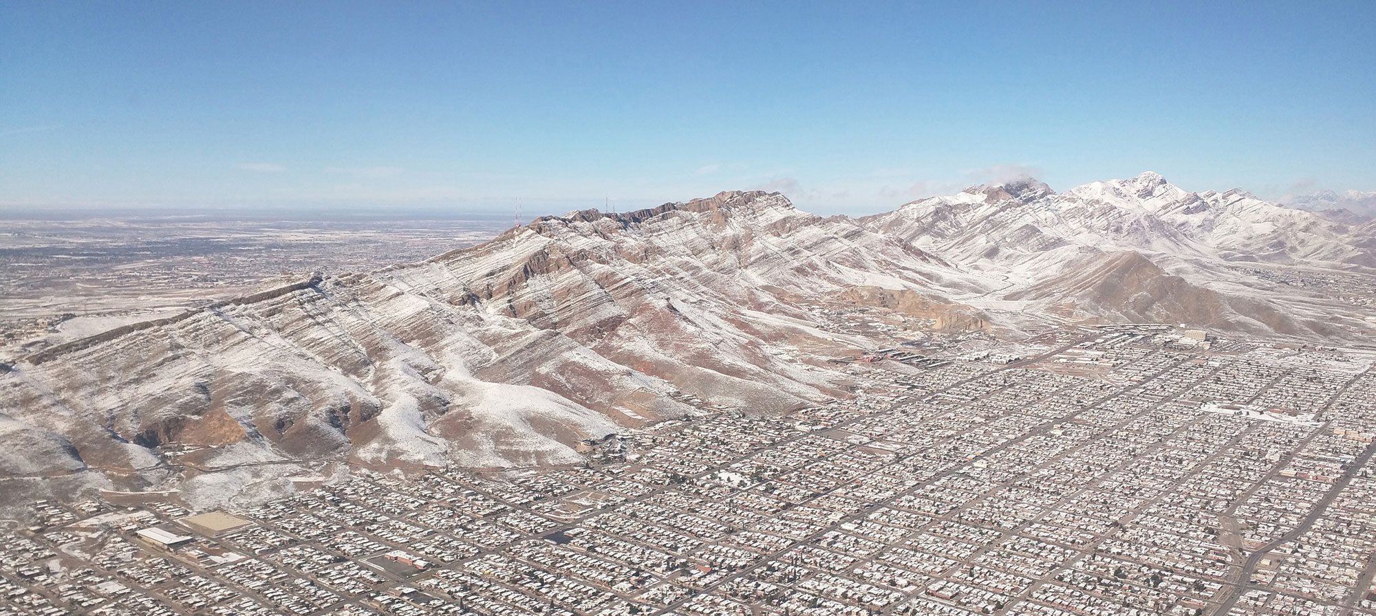 Photograph of the Franklin Mountains in western Texas. The photo shows a mountain range oriented diagonally across the image, with the city of El Paso in the foreground at the foot of the mountains. There is a light dusting of snow.