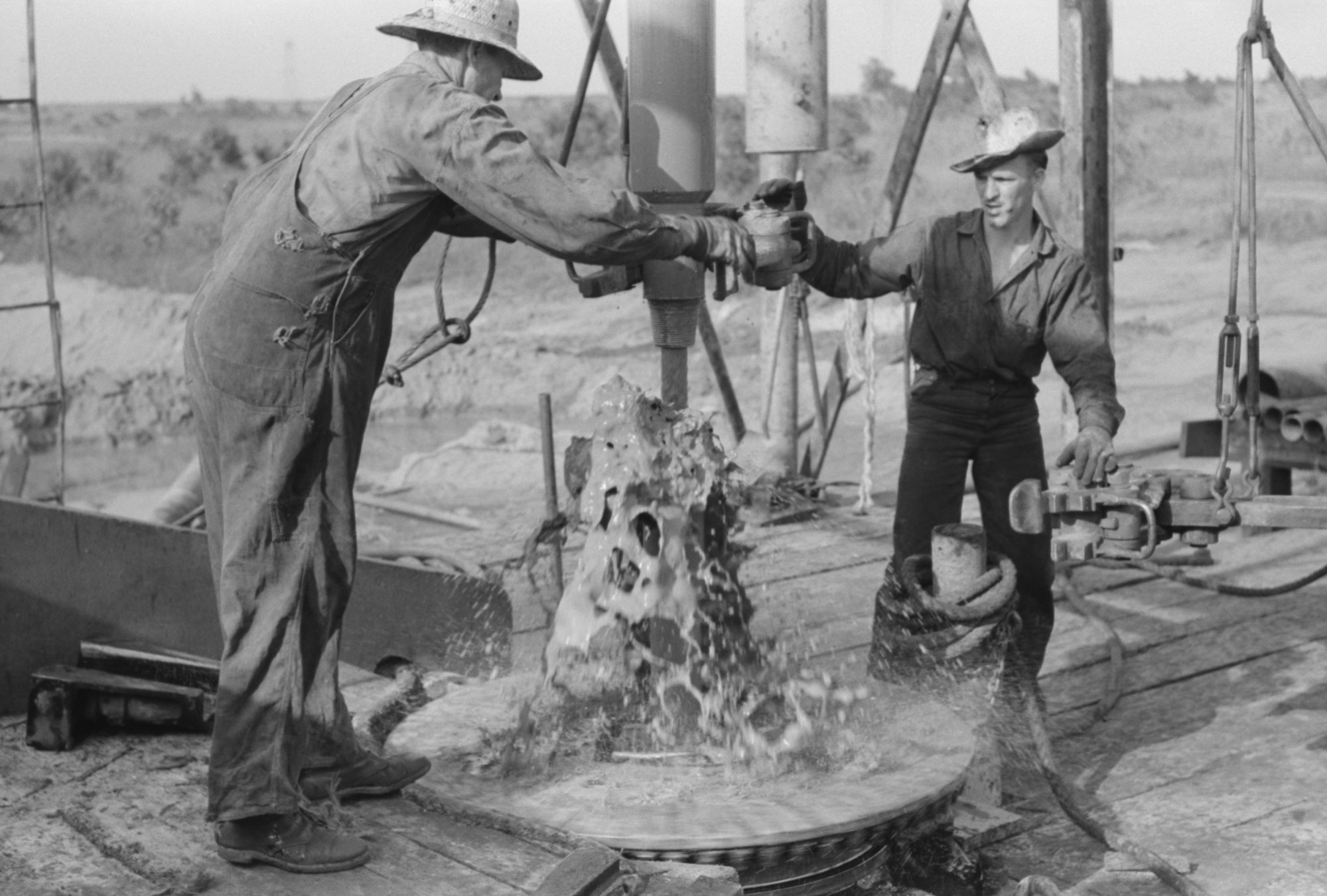 Black-and-white photo of two men adding a length of drilling pipe at oil well. Mud spurts out of the end of the pipe as section of pipes are loosened.