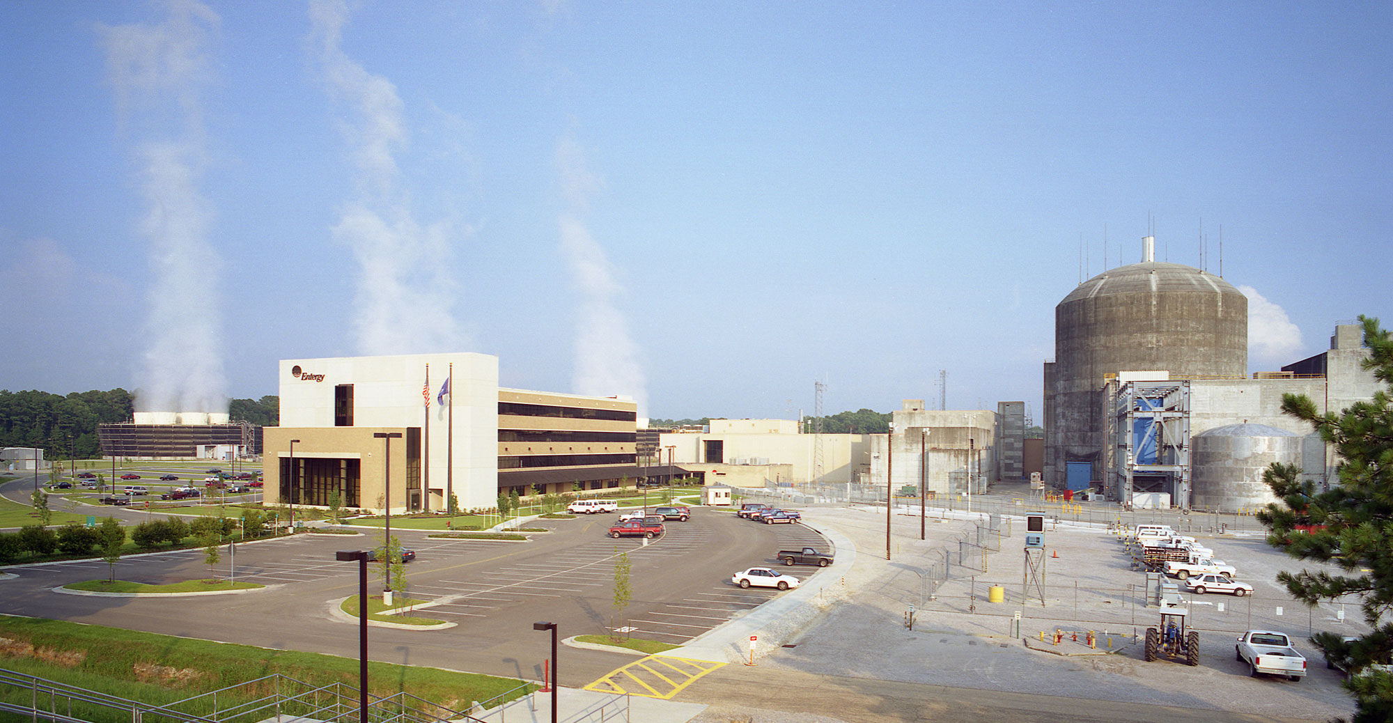 Photograph of River Bend Nuclear Station unit 1, near St. Francisville, Louisiana. An office building and structures associated with the power plant are in the photo. Smoke or steam rises in the background.