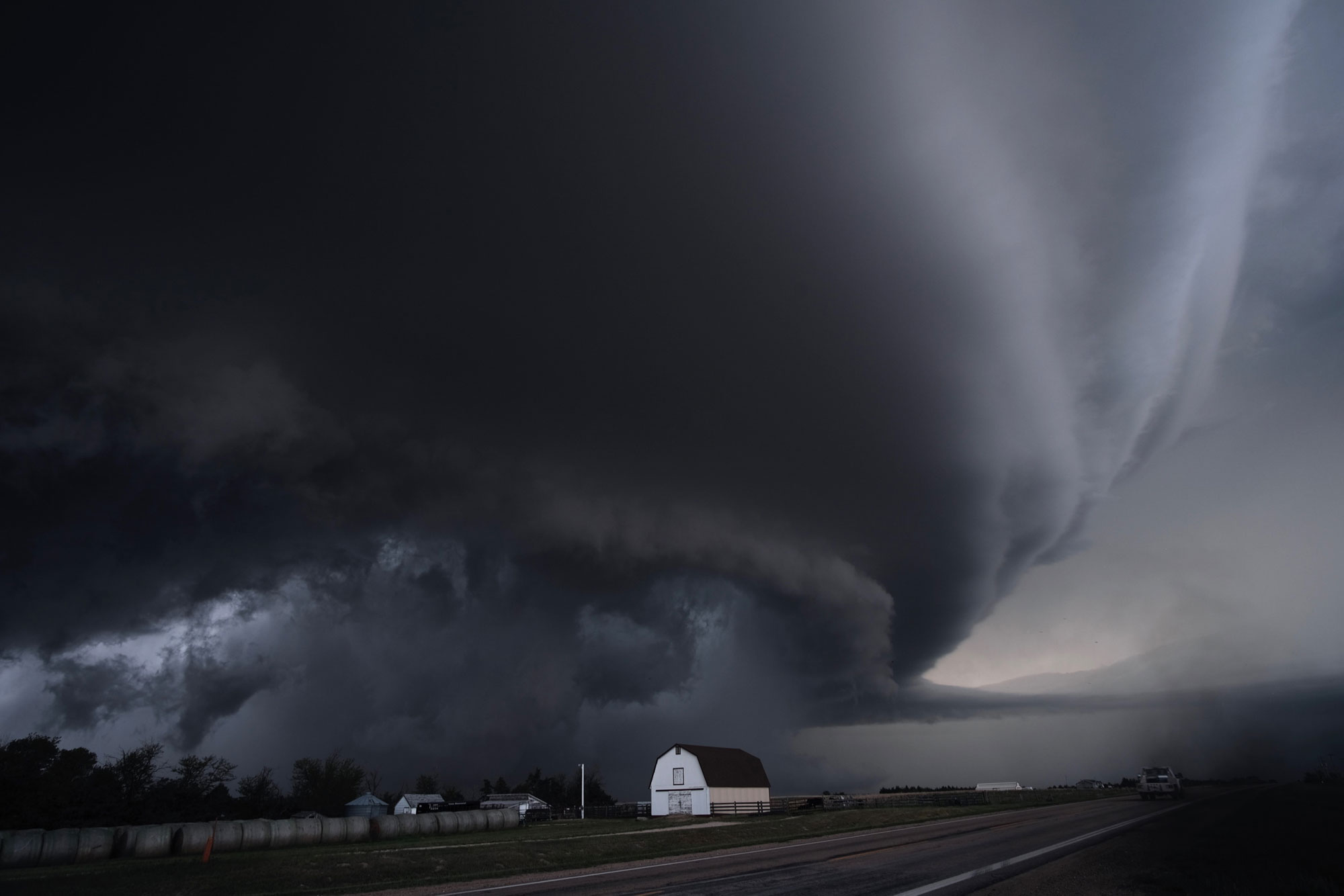 Photo of a supercell storm in Kansas. The photo shows a low-hanging dark cloud over a white barn next to a paved road.