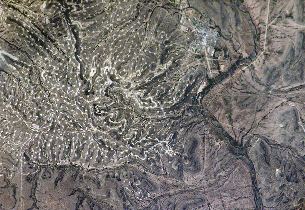 Photograph of the Yates Oil Field, Texas, from taken from space. The photo shows a dry, hilly landscape with sinuous roads and pads for oil wells. The Pecos River is on the right of the image.