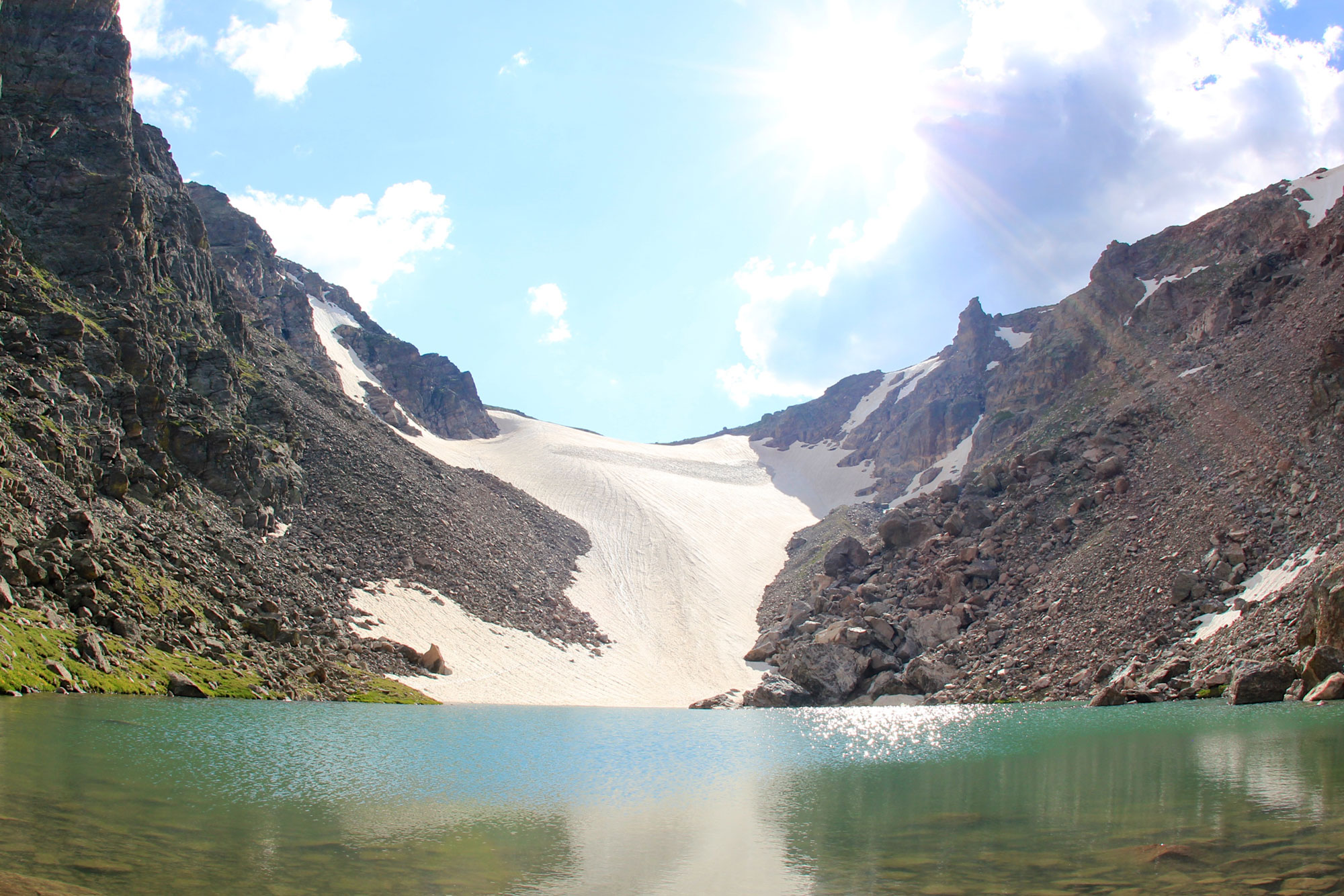 Photograph of Andrews Glacier in Rocky Mountain National Park. The photo shows a saddle between two hills. Snow (the glacier) occurs at the bottom of the saddle and appears to flow downhill, ending at the shore of a lake.