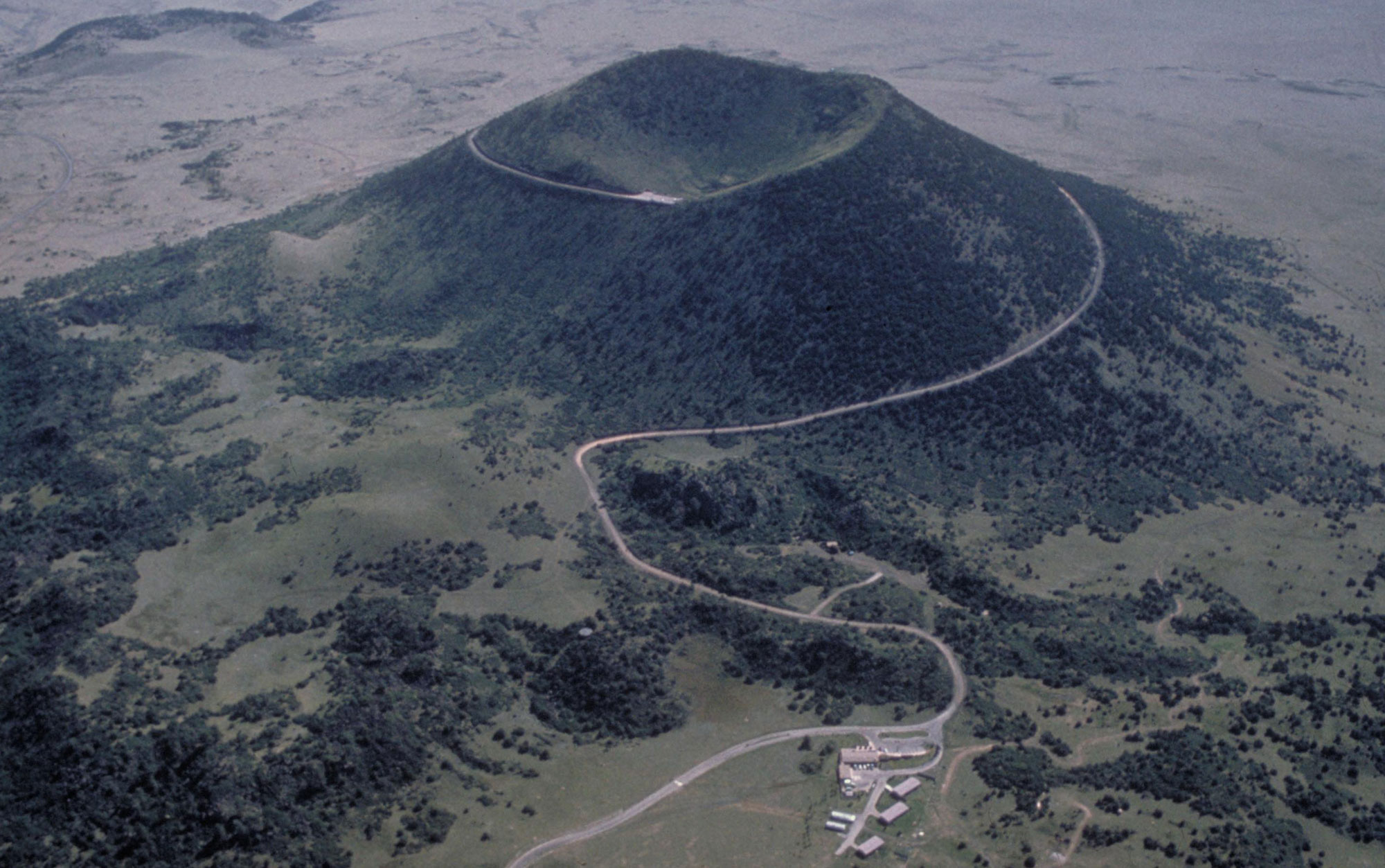 Photograph of Capulin Volcano in northern New Mexico. This volcano is an ancient caldera. It is a cone-shaped hill rising from a flat landscape. The top of the hill has a large depression in it. Building and a parking area can be seen at the base of the hill (bottom or the image). A road spirals up the hill to the top.