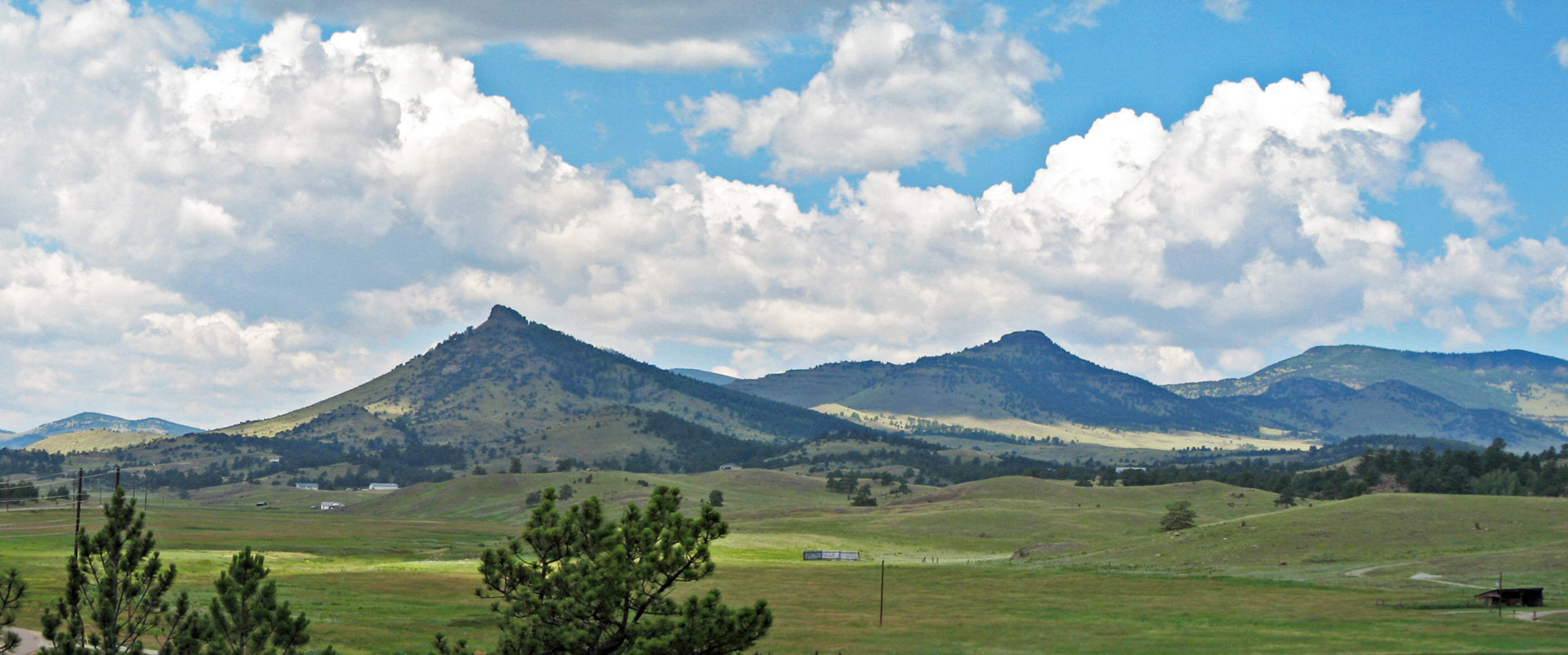 Photograph of the Guffey Volcanic Center volcanoes in Colorado. The photo shows a landscape with a green field in the foreground. Behind it are rolling hills, then taller volcanic peaks. Two of the most prominent peaks are cone-shaped.