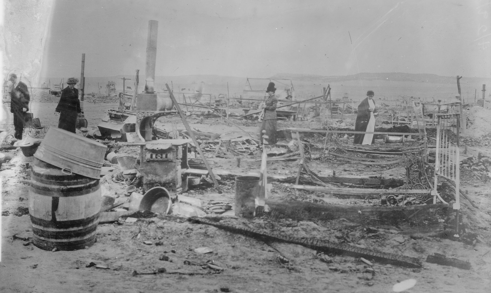 Black and white photo showing the aftermath of the Ludlow massacre. Three women in long dresses and one man stand in a landscape strewn with debris, including a barrel with a metal basin on top in the foreground and a bedframe. What appears to be a chimney is in the background.