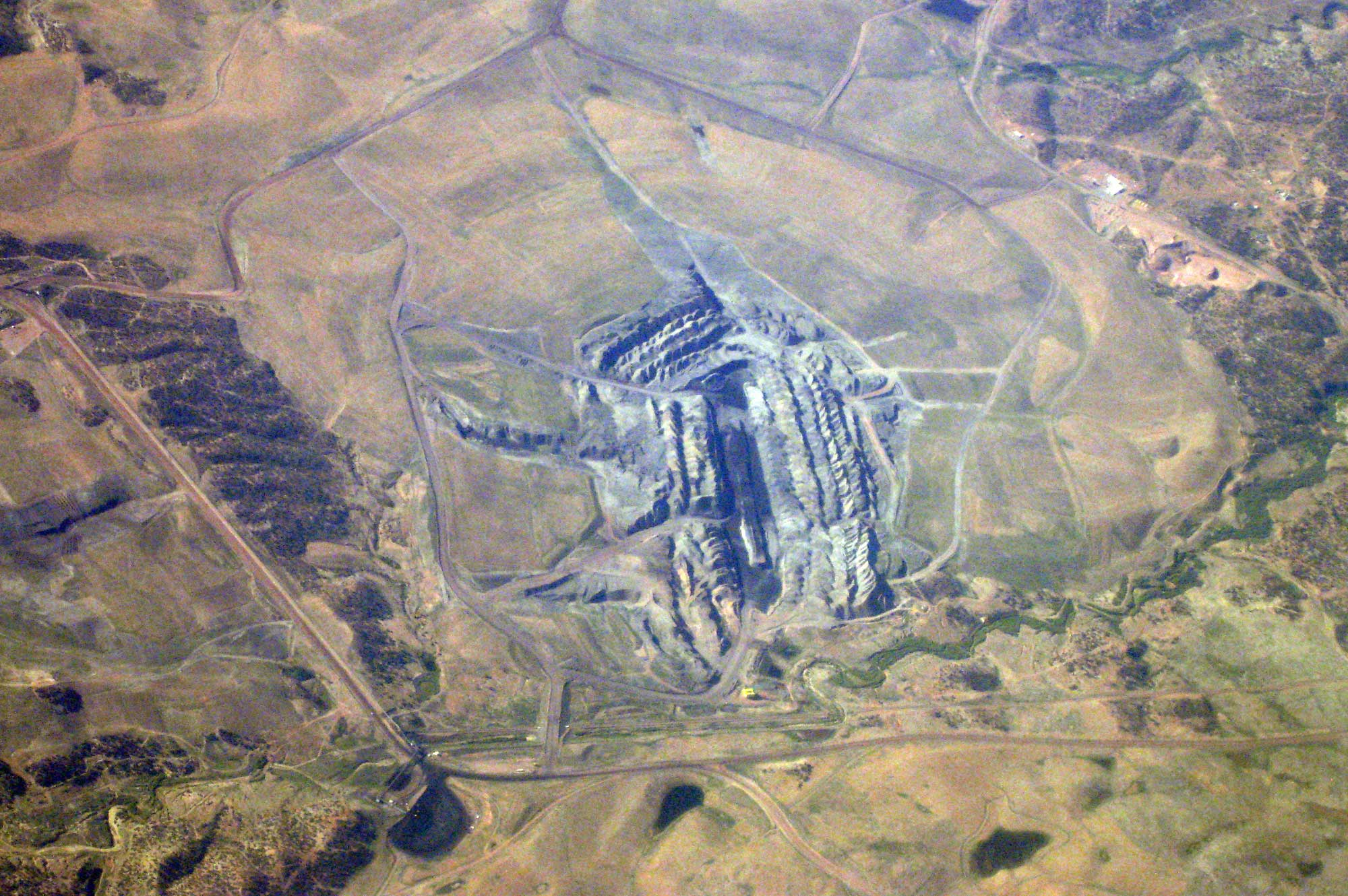 Aerial photo of the Kayenta Coal Mine, a strip mine in Arizona. The Mine looks like an oblong gray landform with a central dark gray region that has been dug into a light greenish-yellow landscape. A road encircles the mine.