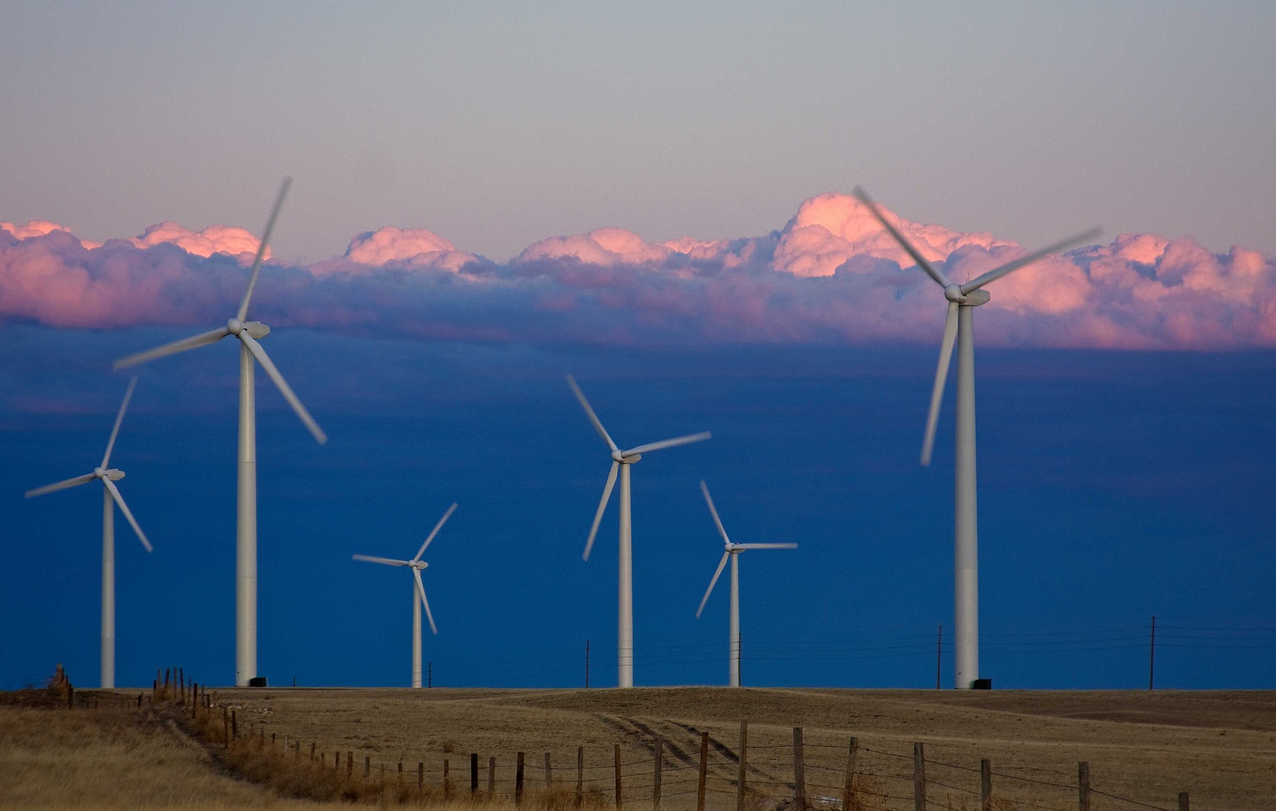 Photo of a windfarm in Colorado. The photo shows six large white wind turbines standing in a field, some in the foreground and some in the background. The sky is cloudy.