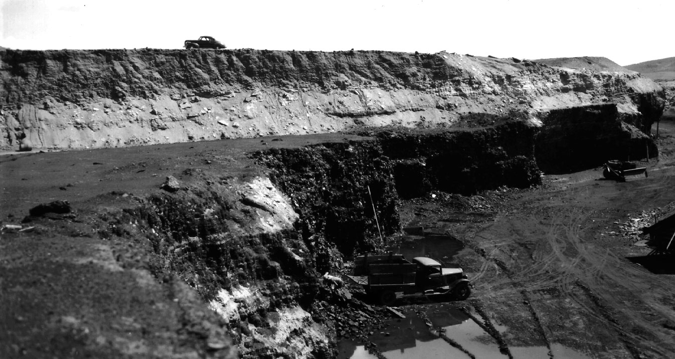 Black-and-white photo of an open-pit coal mine in Colorado. Coal is exposed in an excavated cliff near the floor of the mine, where a truck is being loaded. At surface level above the pit, a car can be seen.