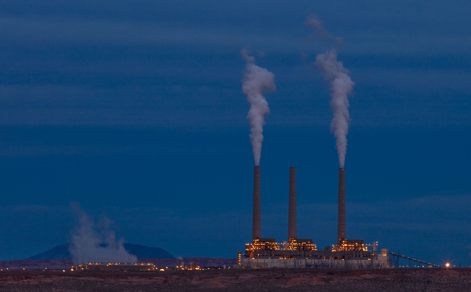 Photograph of a coal-fired powerplant probably taken at dawn or dusk (the sky is relatively dark). The plant has three smokestacks, two of which are emitting smoke. The buildings at the base of the plant have lights illuminated.