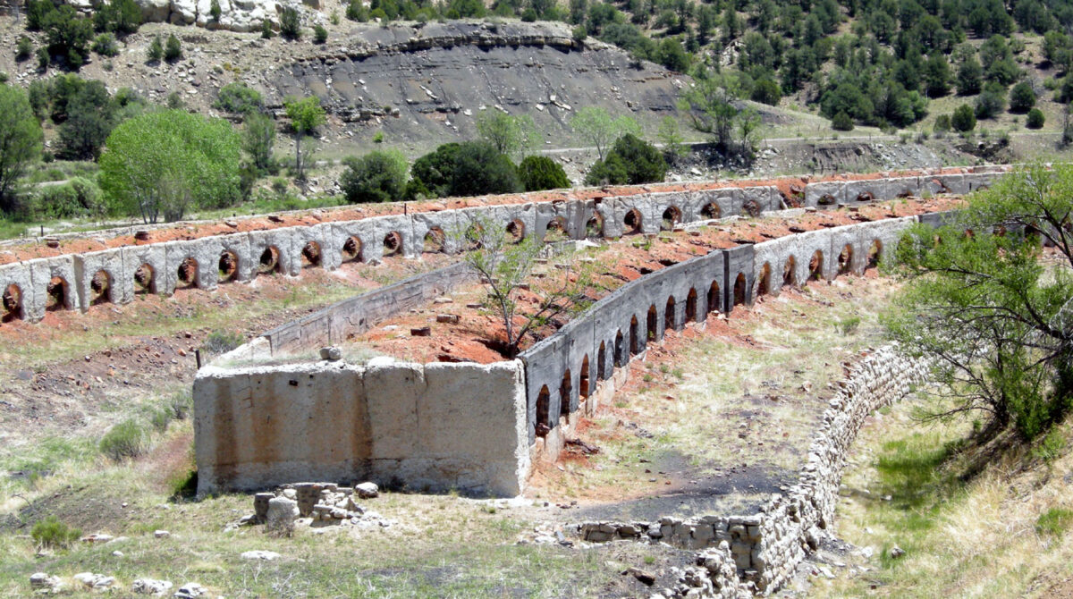 Photograph of abandoned coke ovens in Colorado. The coke ovens may be made of stone or brick. The are elongated structures with multiple arched openings on one side. The opening of two rows of coke ovens can be seen in the image.