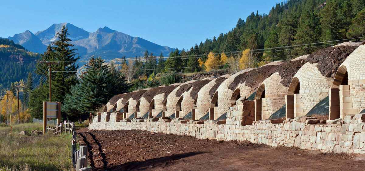 Picture of a row of beehive-style coke ovens, Redstone, Colorado. The coke ovens are made of bricks are are dome- or beehive-shaped. Each has an arched opening. A flat dirt road runs along the length of the row of ovens. A mountain rises in the background.