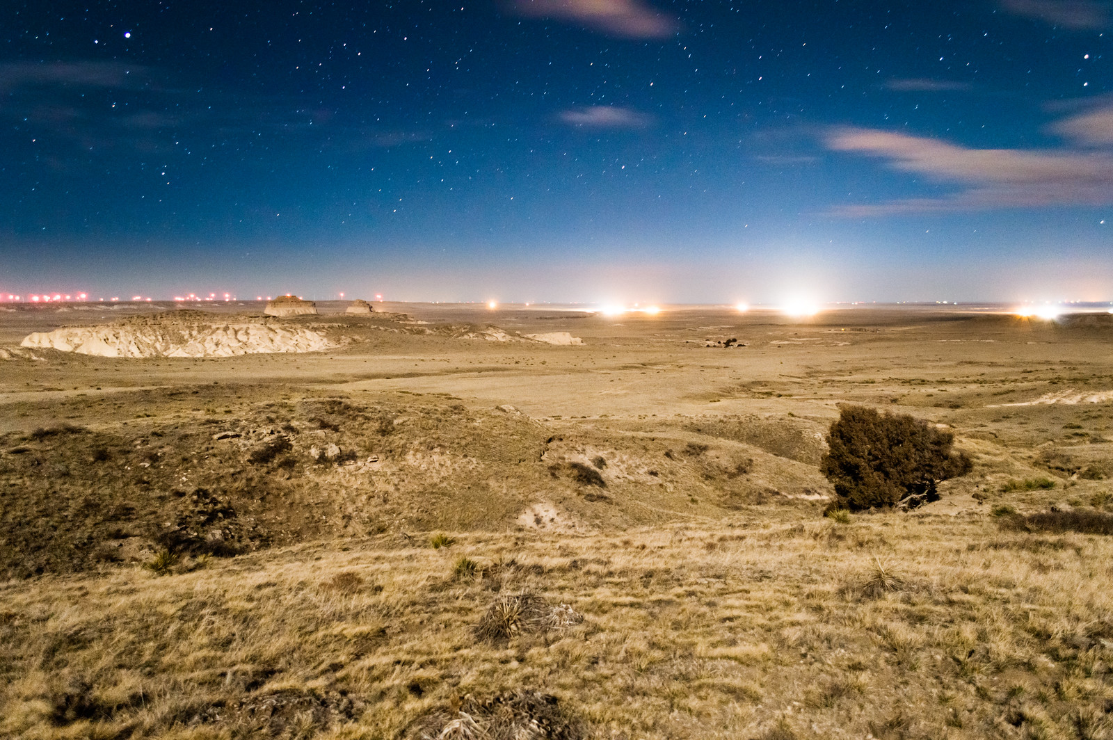 Photograph of a grassy prairie or plain, probably at dusk or at night. Bright lights illuminate the horizon. On the left, lights are mostly small and orange in color. On the right, the lights are much brighter and white in color.