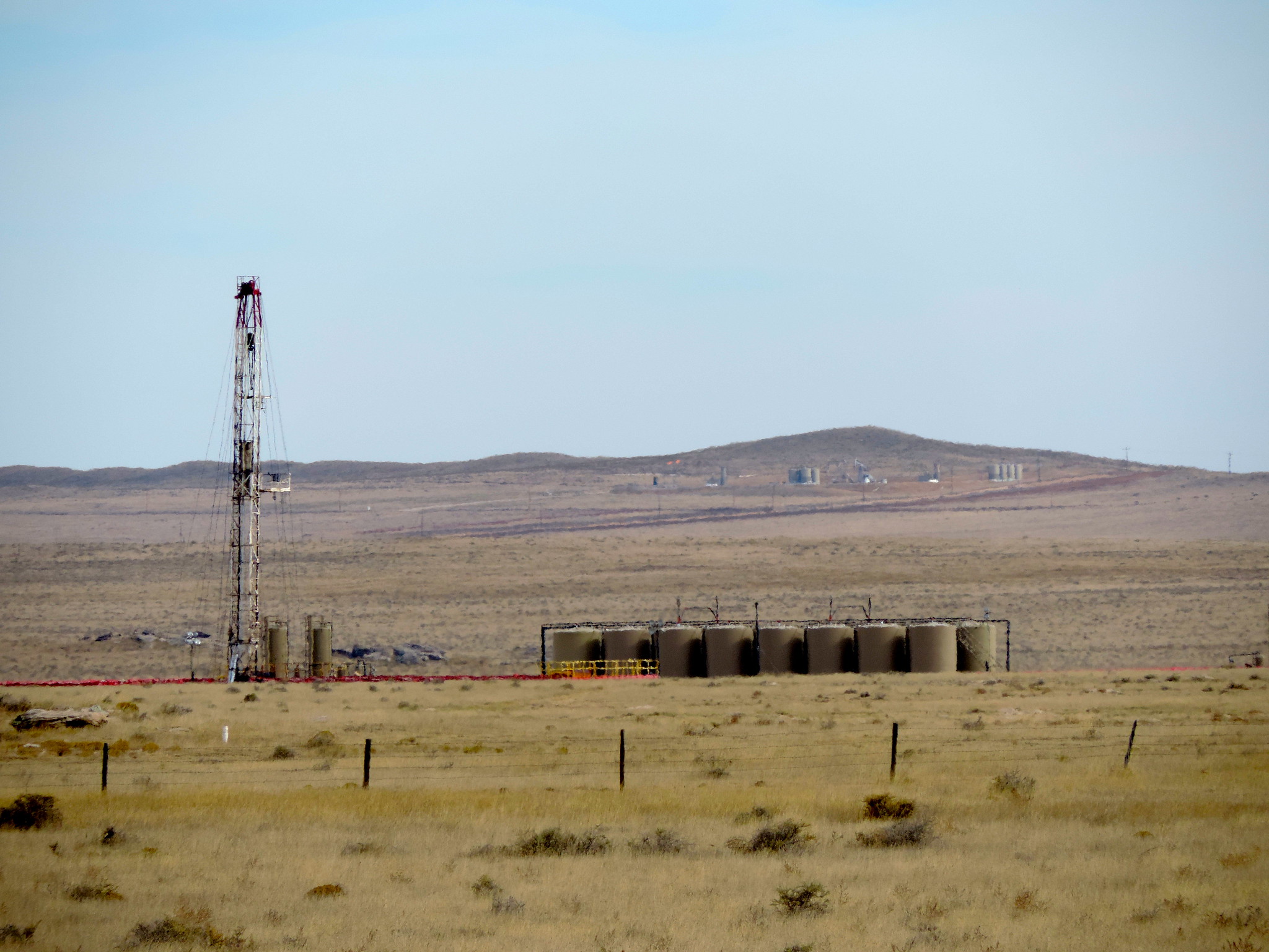 Photograph of a drill right (a metal tower) and a battery of oil tanks on a relatively flat grassland. In the background, a low hill with what appear to be more oil tanks on its slopes rises.