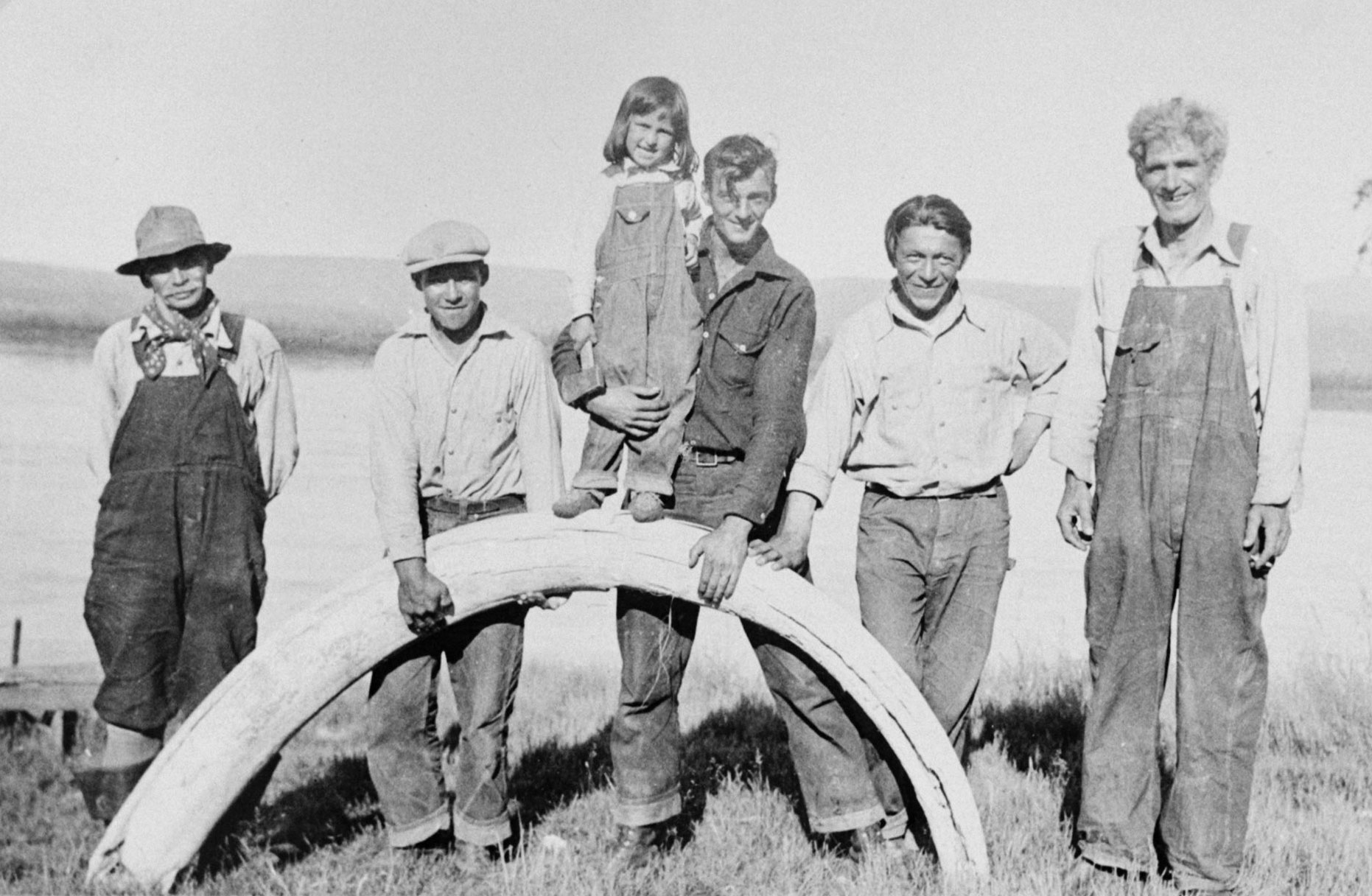 Black and white photo of five men posting with a mammoth tusk. Two of the men are holding up the tusk, which has its base and tip resting on the ground. The man in the center holds a little girl who is standing on the apex of the arch formed by the tusk. 