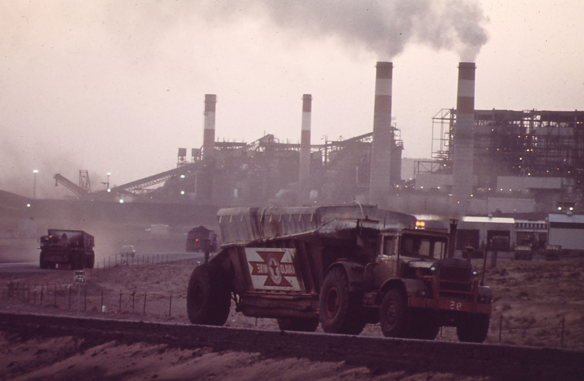 Photo showing four smokestacks and other structures of the Four Corners Generating Plant in the background, with trucks transporting coal to the plant and an empty truck driving away. The whole scene appears smoky from the plant emissions.