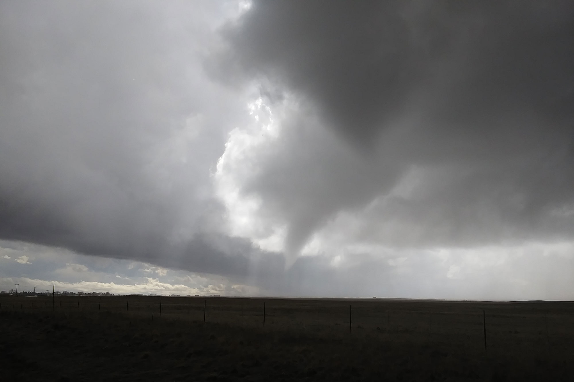 Photograph of a cloudy sky with two funnel clouds (cone-shaped extensions of the clouds) extending towards the ground. On the ground, the landscape is so dark it is nearly black.