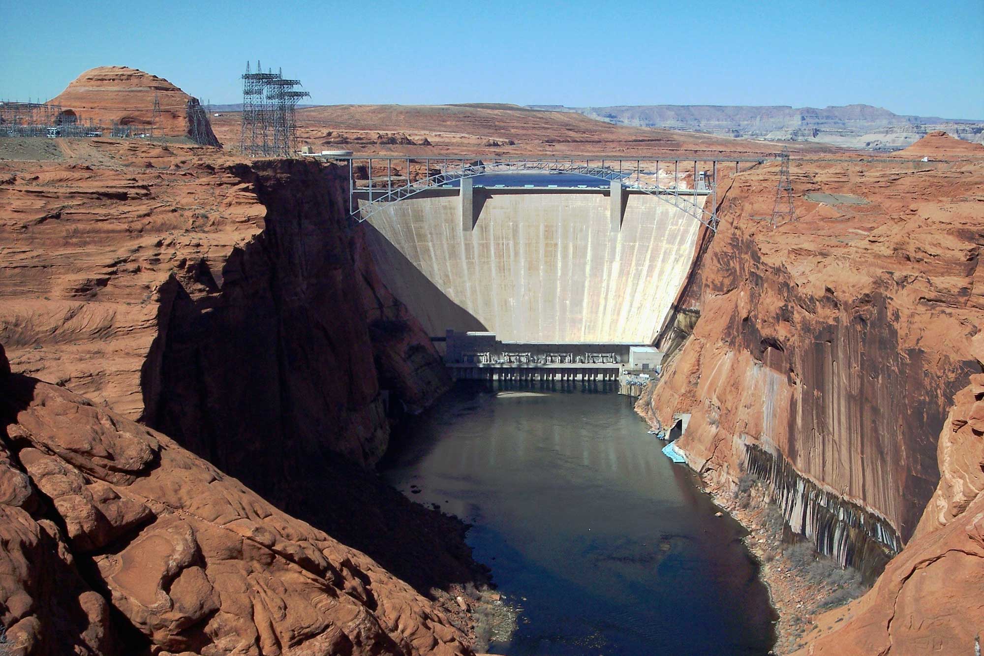 Photograph of Glen Canyon Dam in Arizona. The dam is a tall concrete structure spanning a canyon made up of red rocks. The lake behind the dam can barely be seen. In front of the dam, water level is low. A bridge spans the canyon in front of the dam.