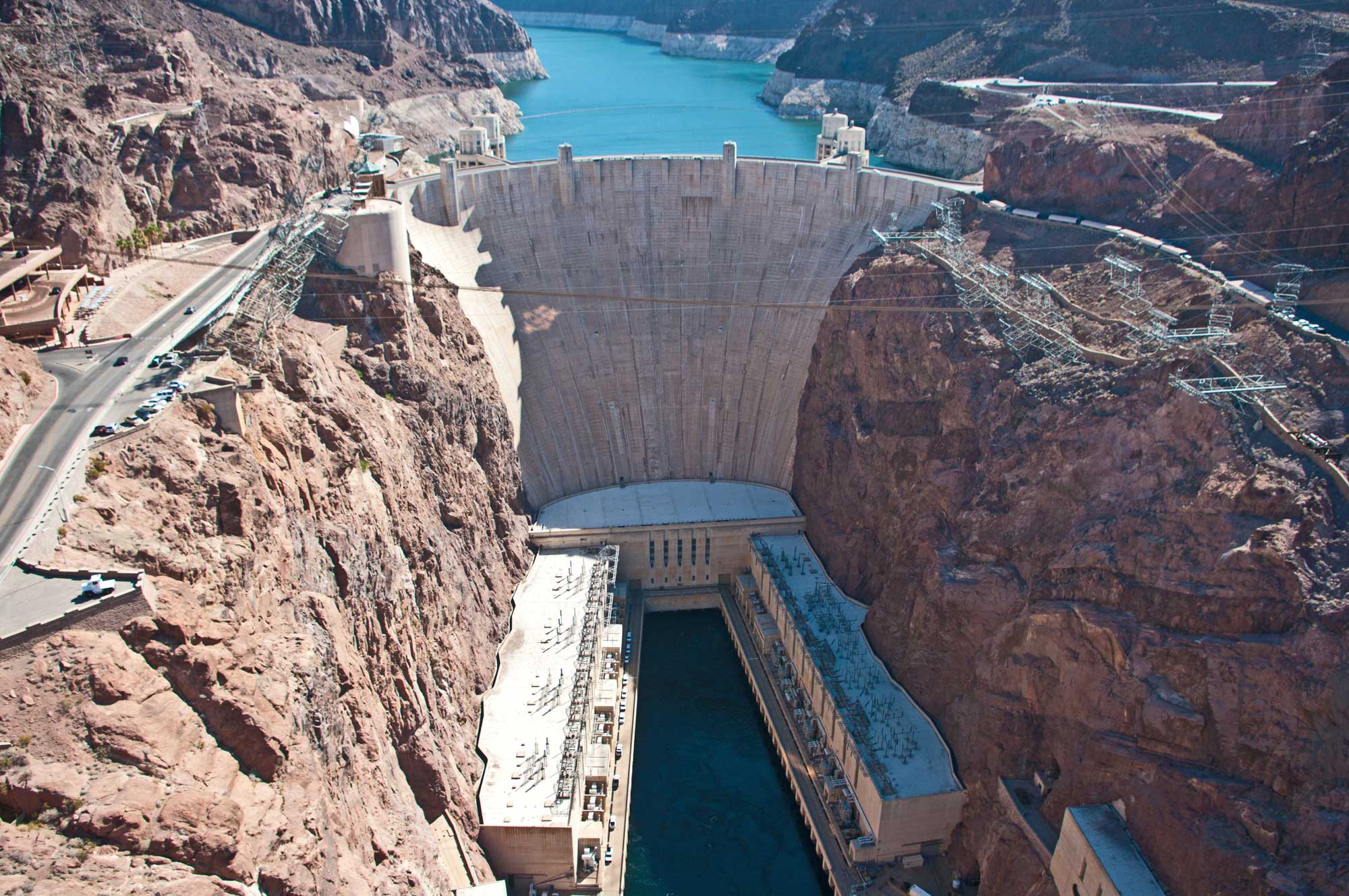 Photograph of Hoover Dam on the Arizona-Nevada border. The dam is a large cement wall holding back water upriver. Downriver (foreground), the water is much lower and hydroelectric structures can be seen. The rocks surrounding the river in front of the dam form red cliffs.
