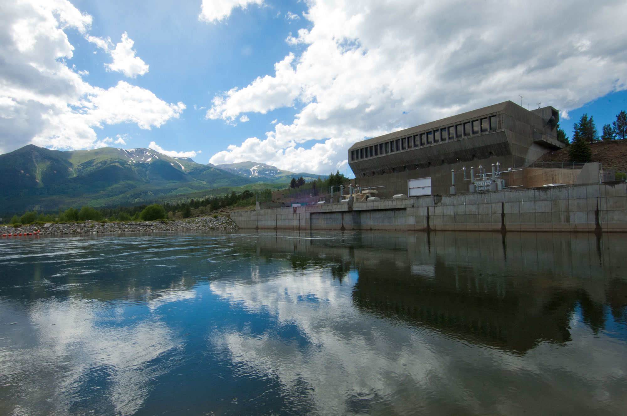 Photograph of a lake with a pumping station next to it. The pumping station is a concrete building with a bank of windows facing the lake. Some equipment sits in the front of the building. Mountain peaks rise in the background.