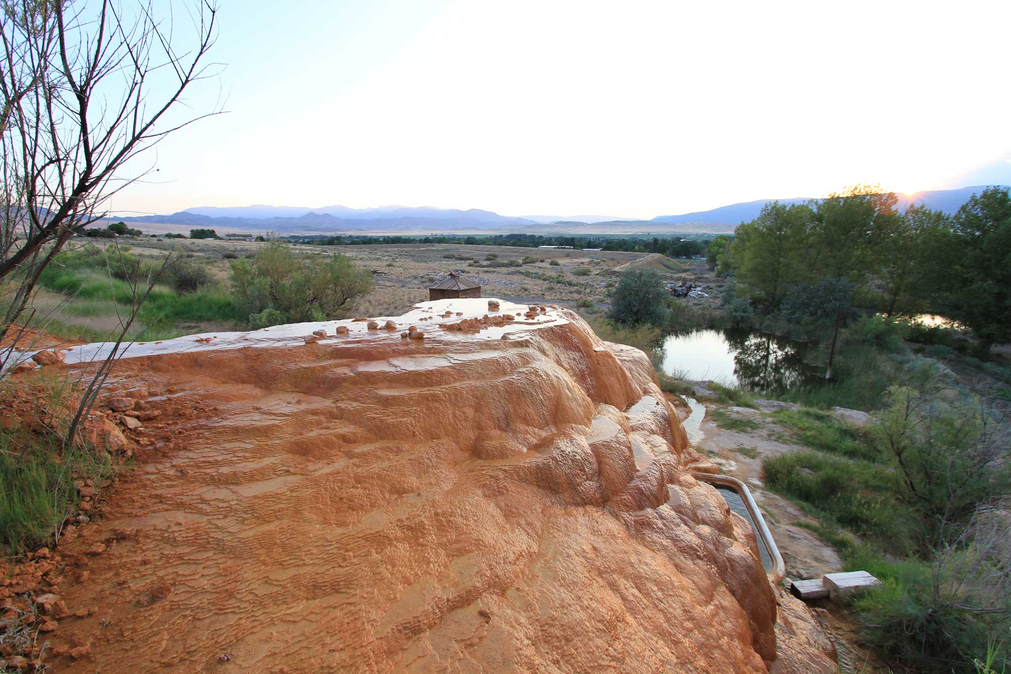 Photograph of Mystic Hot Springs in Monroe, Utah. The hot spring looks like a wet bound of orange rock with a thin film of water sitting on the top. At the bottom edge of the hot spring, a bathtub filled with water can be seen. In the background is a dry landscape with a pool of water. Mountains are on the horizon.