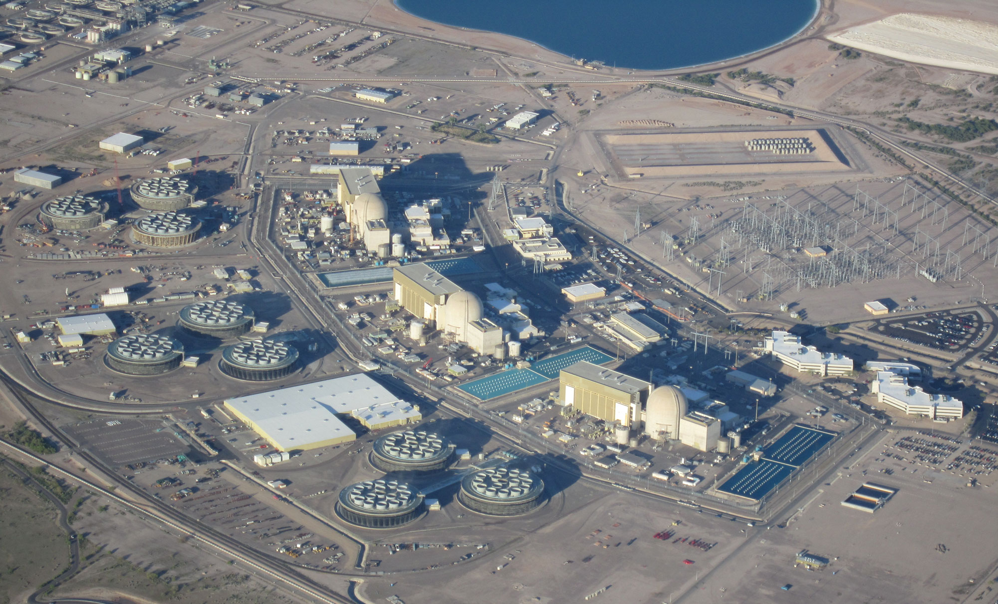 Aerial photo of Palo Verde Nuclear Generating Station in Arizona. The photo shows three reactors and associated building and infrastructure on a barren landscape. The corner of a lake is the top of the image.