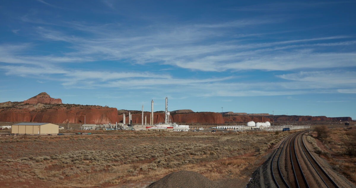 Photograph of the structures of an oil refinery in New Mexico on a flat landscape with low vegetation. Behind the refinery are low cliffs made up of red rocks. The sky is blue with wispy clouds.