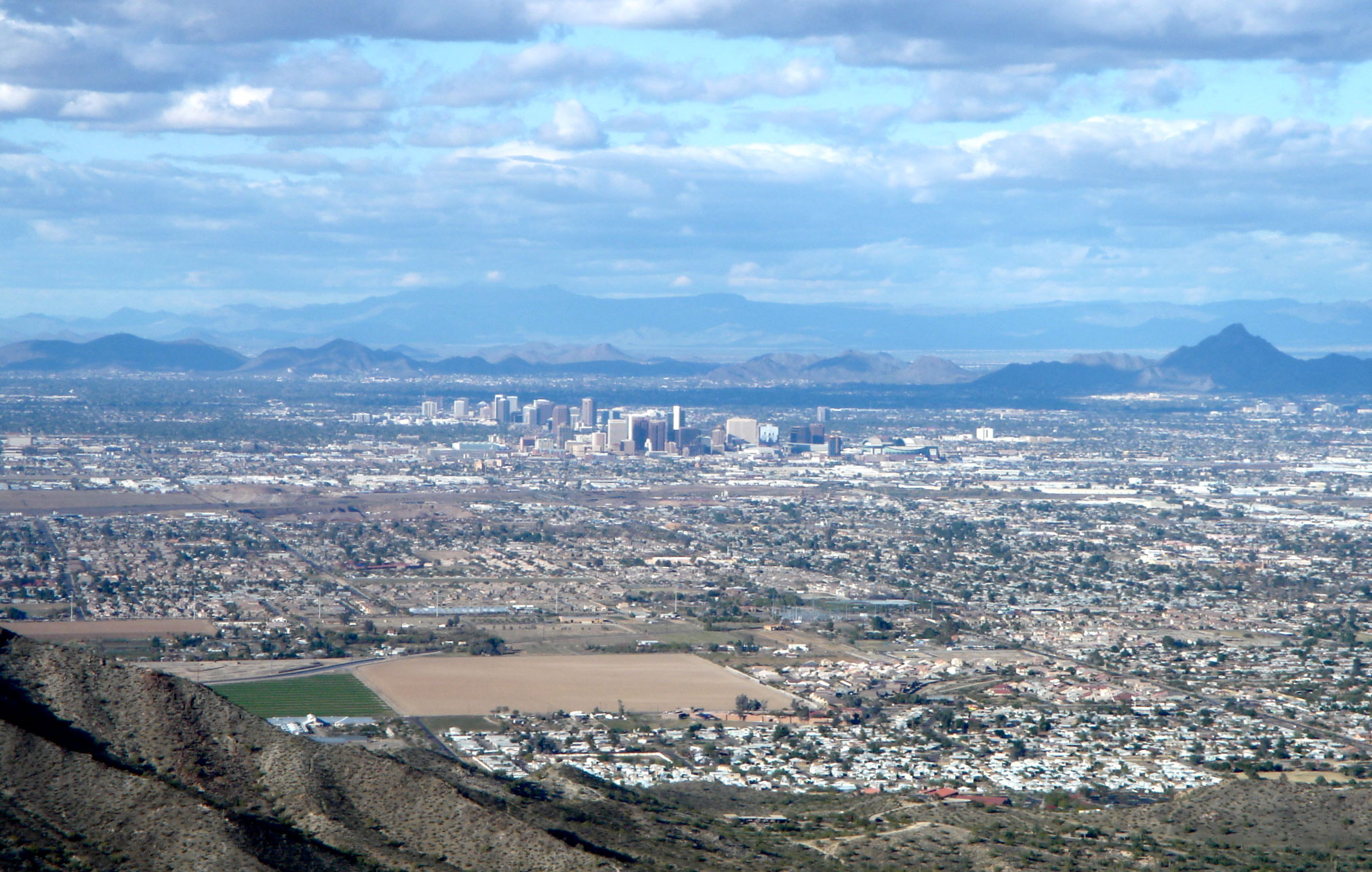 Photograph of Phoenix, Arizona, from a high point near the city. The photo shows development (roads, buildings) on a flat plain with mountains rising in the distance. Between the mountains and the nearest buildings is a cluster of tall buildings in downtown Phoenix.