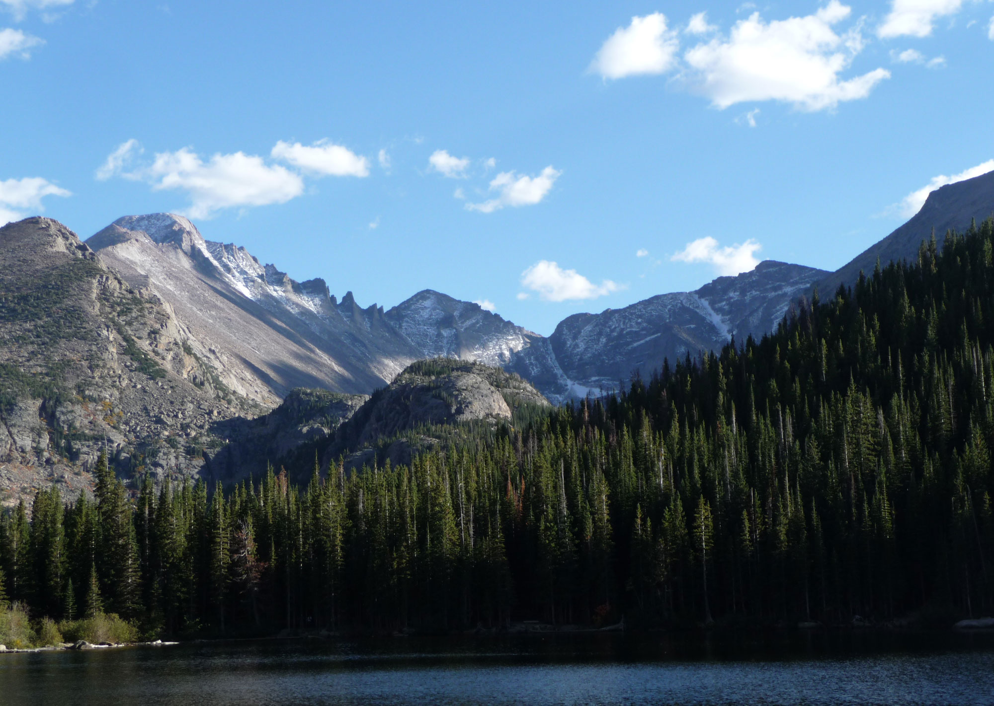Photograph of a lake surrounded by mountain peaks in Rocky Mountain National Park, Colorado. The nearest slope is covered with conifers. More distant peaks have no vegetation and a light dusting of snow. In the foreground is the surface of a lake.