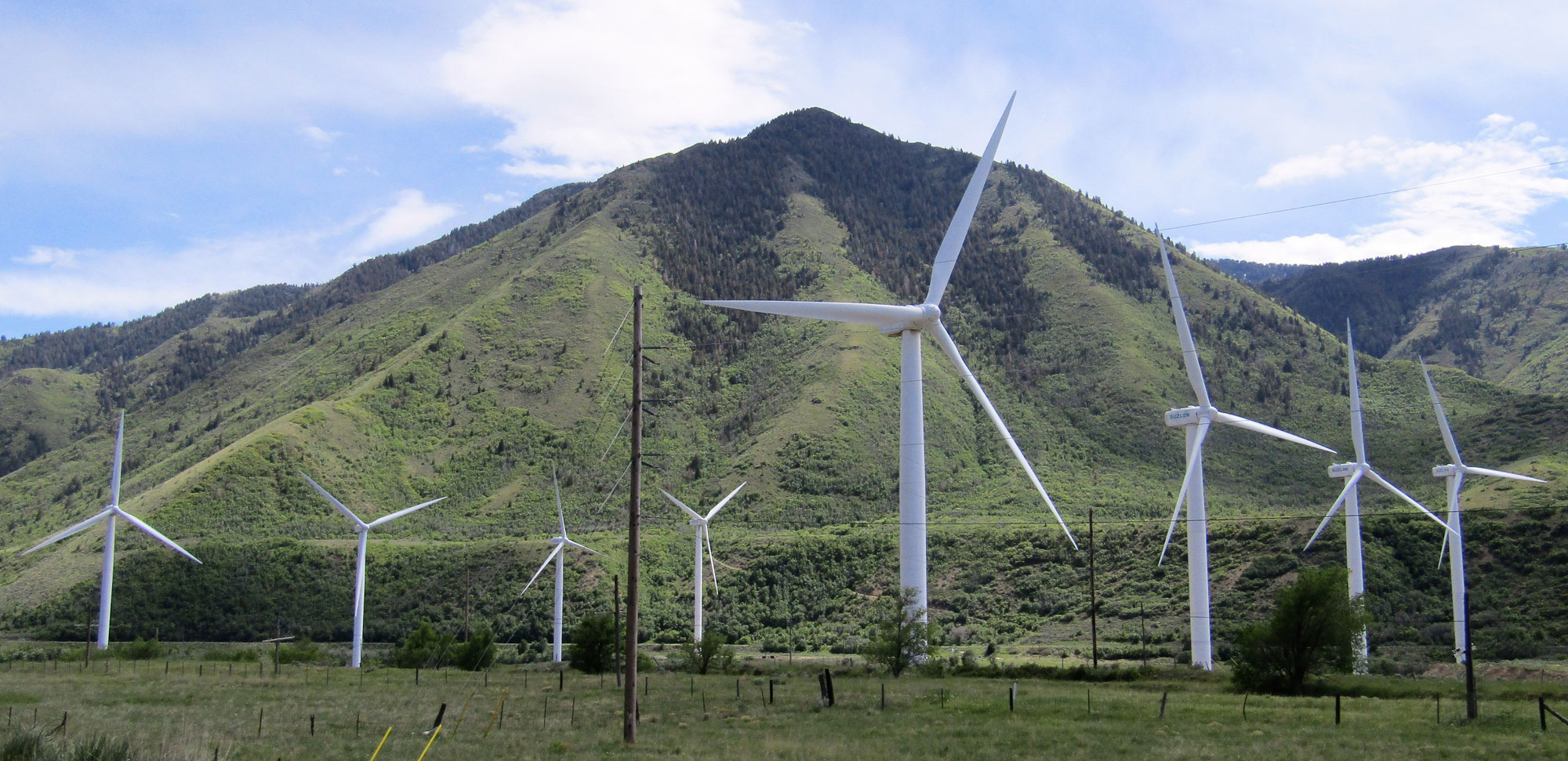 Photograph of a wind farm in Utah. The photo shows eight white wind turbines in a flat field. In the background, a hills rises up being them. The upper summit of the hill is topped with trees, whereas the lower slopes look scrubby.