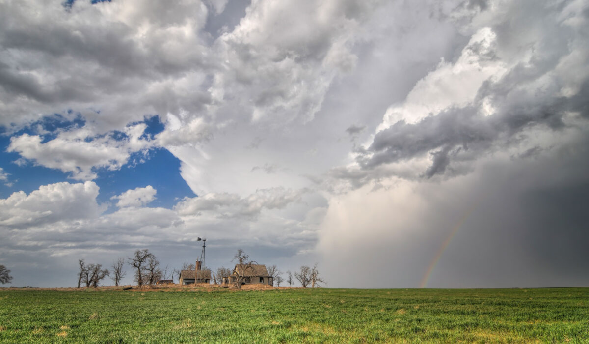 Photograph of a field with buildings and an old windmill surrounded by trees without leaves on the horizon. The sky is partly cloudy to the left, stormy with a rainbow to the right.