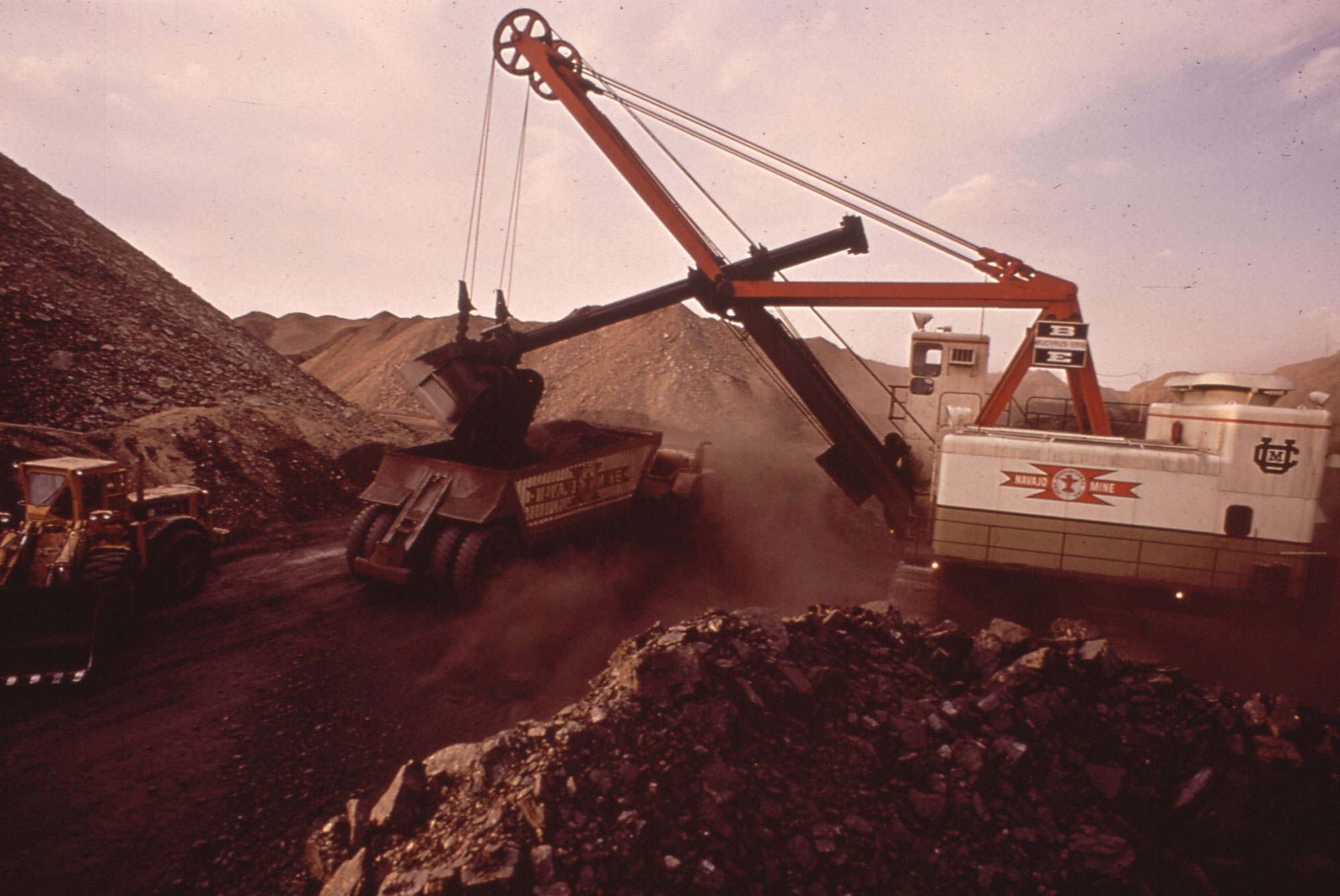 Photo of a strip mining operation in New Mexico. A large shovel operated by a pulley dumps a load of coal into the back of a waiting truck. Mounds of coal can be seen in the foreground and background.
