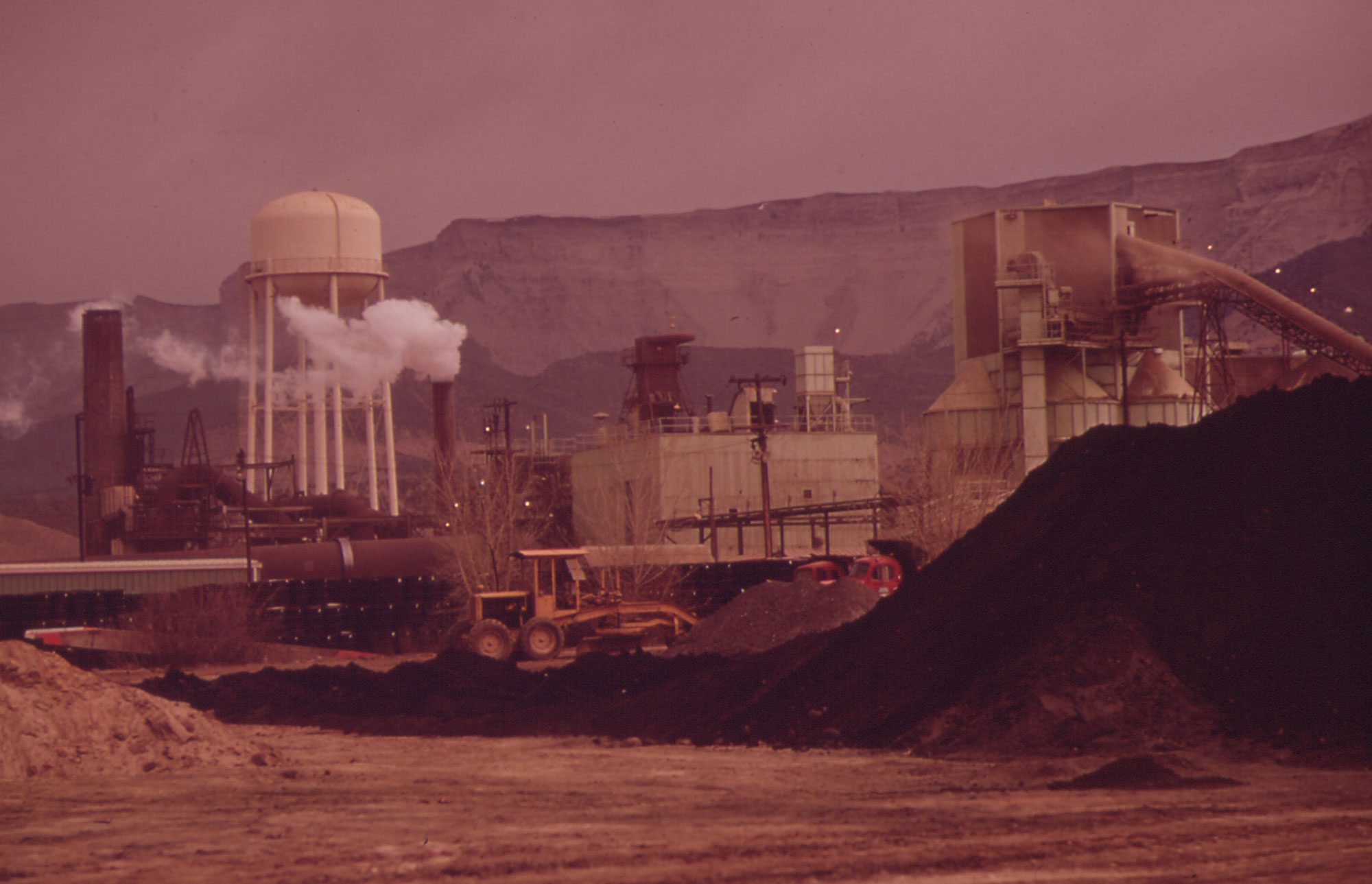 Photograph of a uranium mill in Colorado, 1972. The mill includes a water tower, smokestack, and other structures. In the foreground of the image is a mound of black rock of unknown identity. Tall cliffs rise behind the mill in the background.