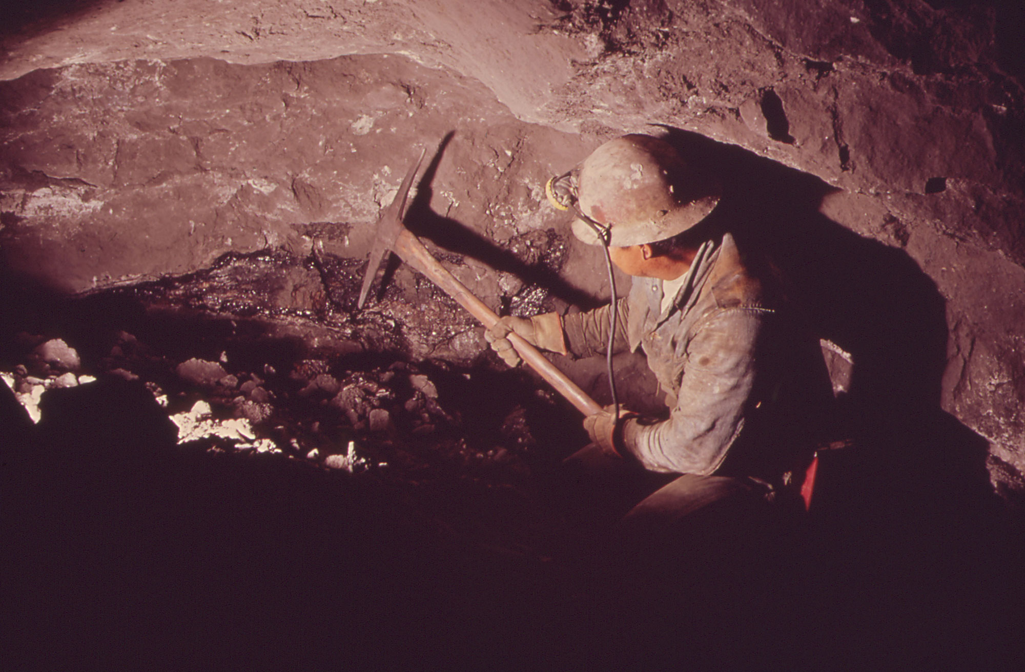 Photograph of a miner in an underground uranium mine. The miner wears a hardhat with a light on the front and holds a large pick in both hands. He is dressed in a long-sleeve blue button-down shirt and wears leather gloves. The rock surrounding him is gray.