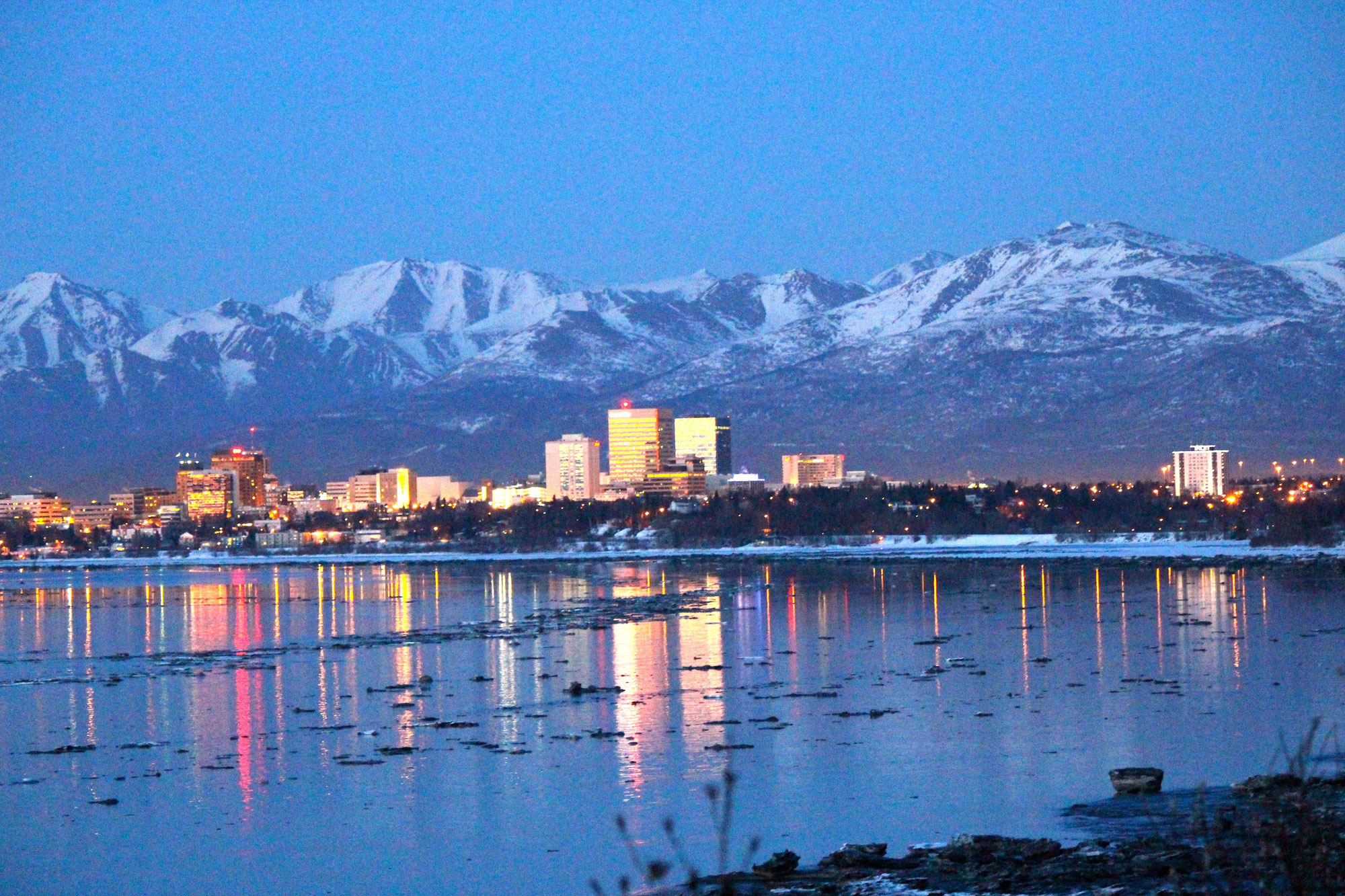 Photograph of Anchorage, Alaska, taken in 2016. The photo shows a city along a coast with snow-capped mountains rising in the background. The photo was taken in semi-darkness and the city is illuminated by lights.