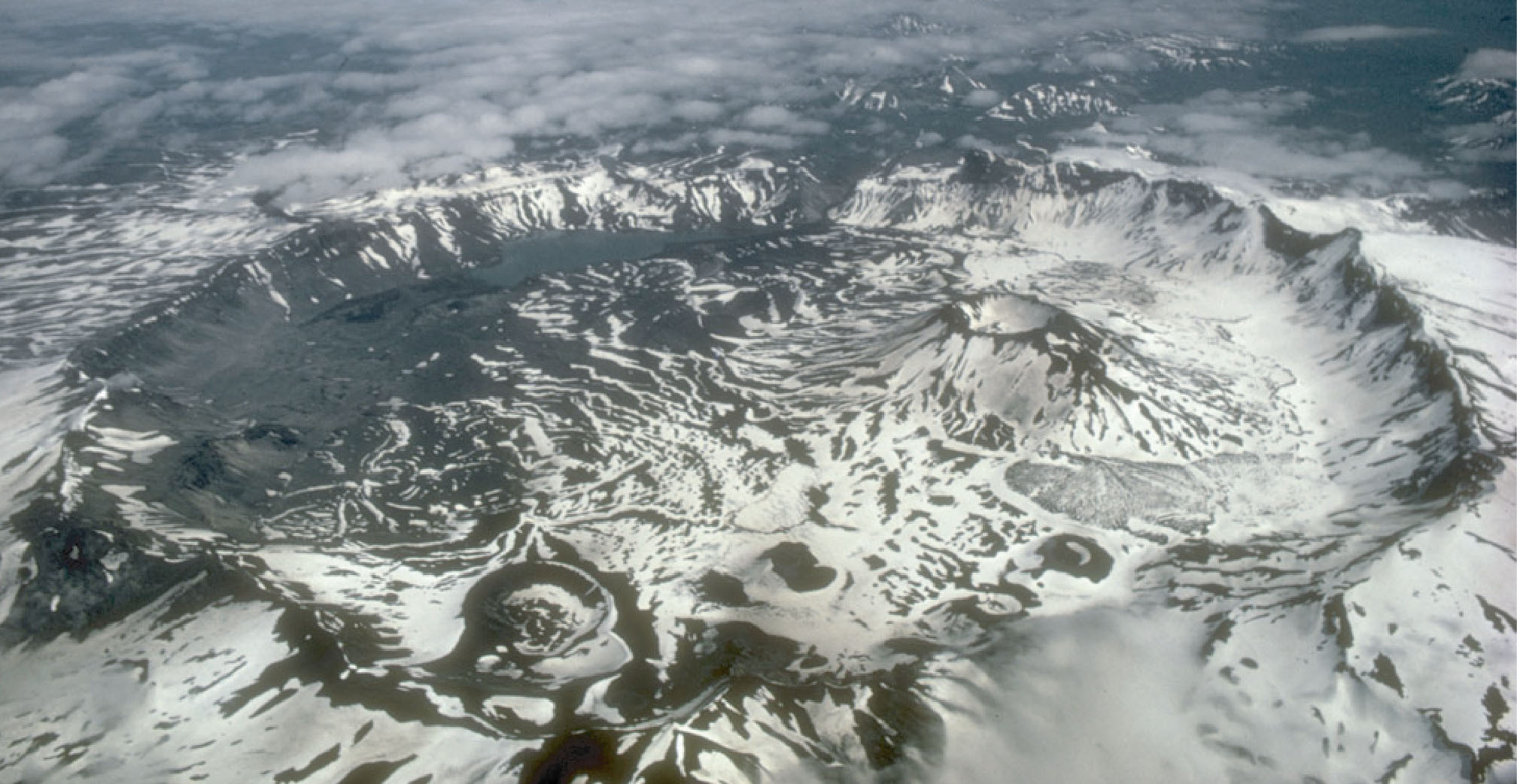 Aerial photo of Aniakchak caldera in Alaska. The photo shows a circular depression in the landscape surrounded by a raised rim. Inside is a projecting volcanic cone. The landscape is brown and dusted with white snow.