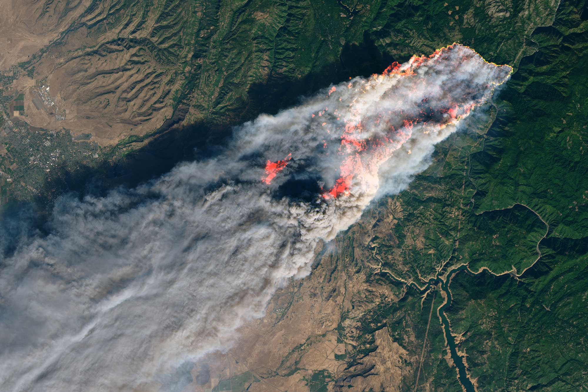 Satellite image showing a plume of smoke on a rugged landscape around Paradise, California, in November 2018. The smoke runs from the upper right to the lower left of the image. In the center of the image, orange areas can be see within the smoke plume. These are areas that are burning as indicated by viewing them in infrared light.