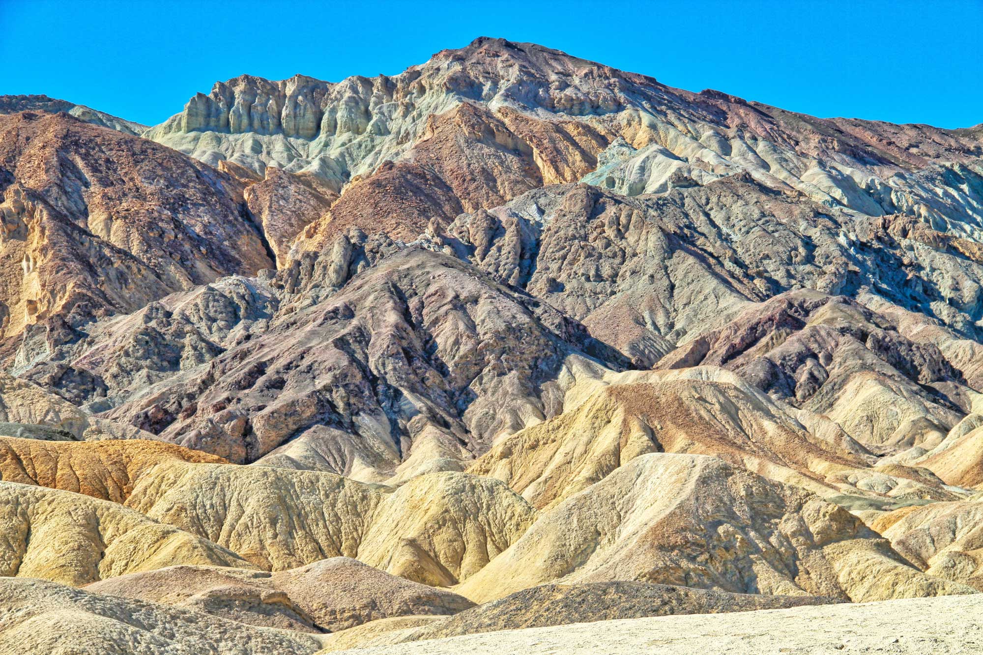 Photograph of the Artist's Palette in Death Valley. The photo shows a landscape of badlands with no visible vegetation. Badlands range in color yellowish to slightly pinkish to orange to greenish.