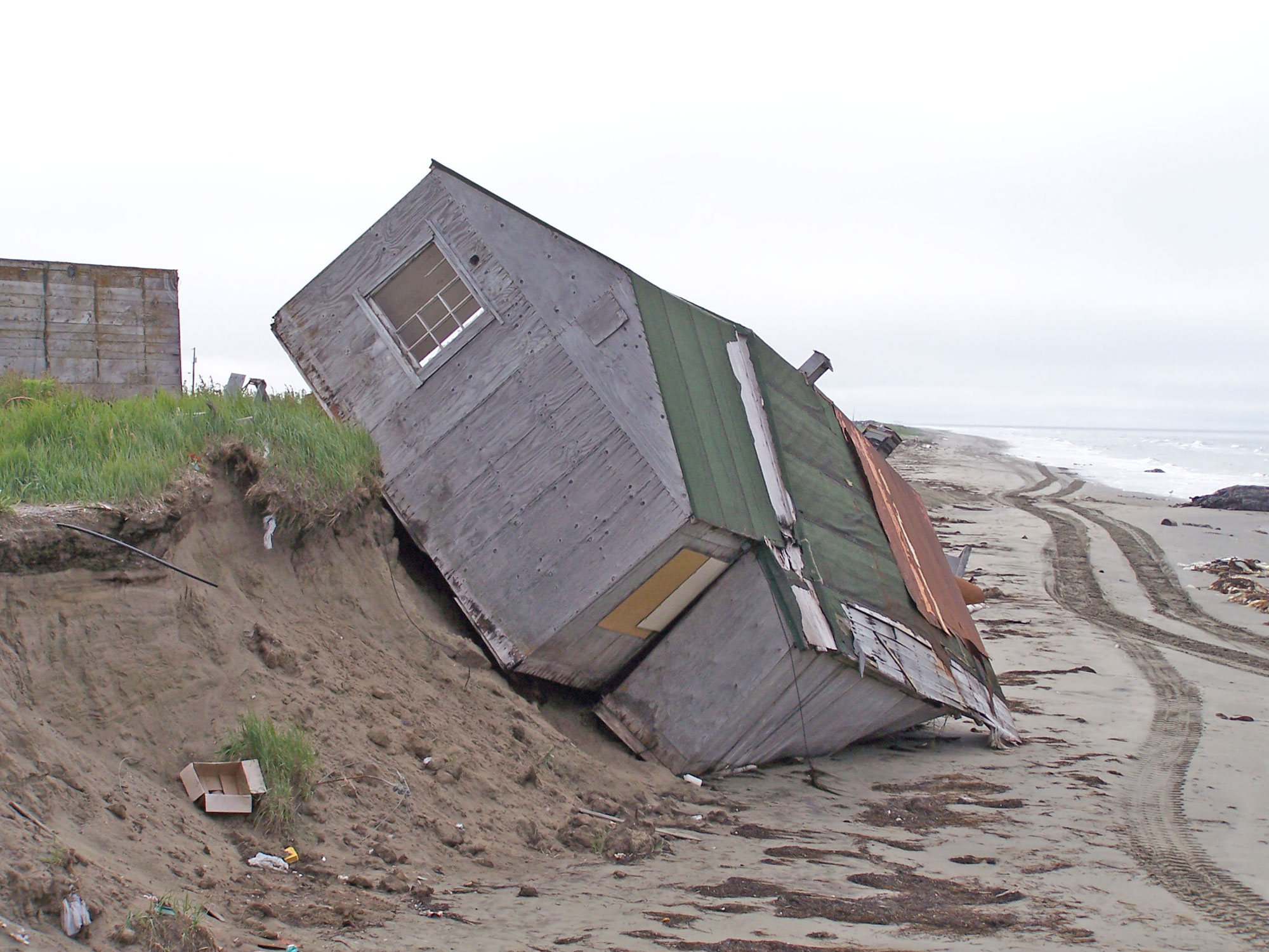 Photograph of a home that has been undermined due to coastal erosion on the Seward Peninsula in Alaska. The photo shows a home sided with unpainted wood, that is tilted over the edge of a sandy coast bluff. One edge of the home is on resting on the beach, while the upper edge is lifted off the ground. 