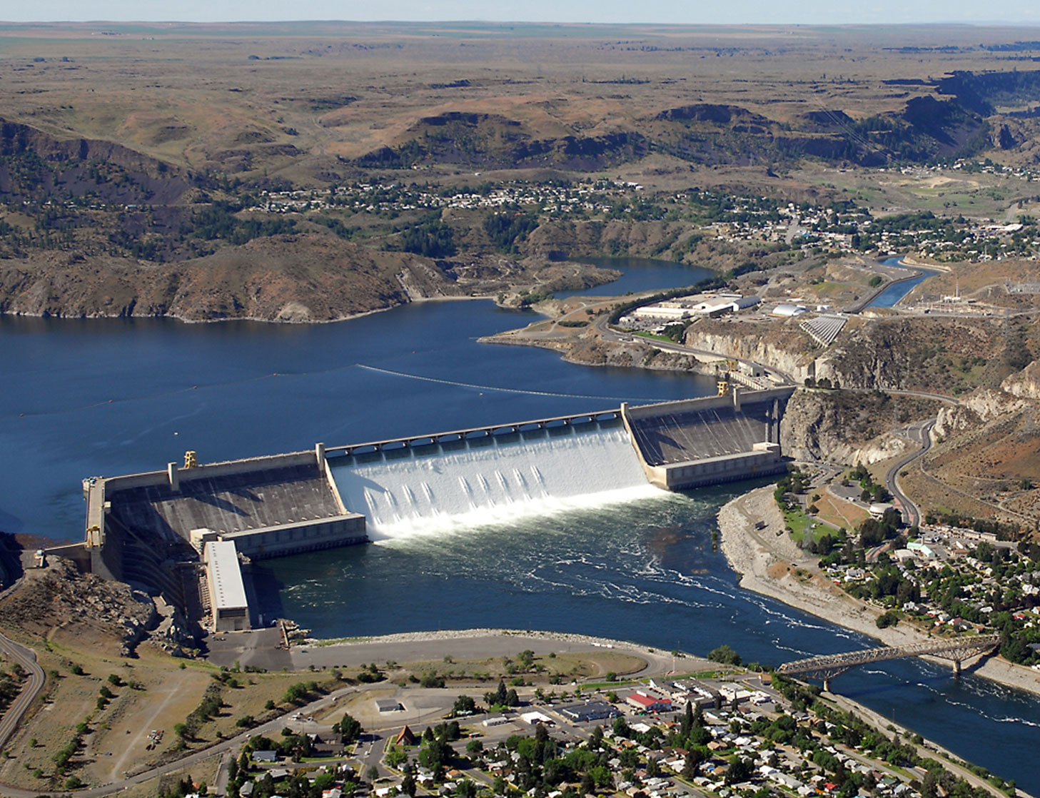 Photograph of Grand Coulee Dam on the Columbia River in Washington state. The dam is a concrete structure holding back water in a reservoir. Water is flowering over the spillway into the river below. The surrounding landscape is dry with low hills.
