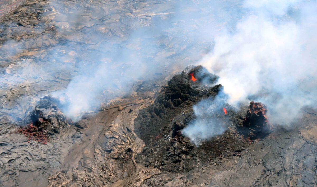 Photograph of vent cones at Halema'uma'u Crater on Kilauea, Hawaii, in 2022. The photo shows a cluster of three cone-shaped structure and a single cone-shaped structure to the left, all of them venting smoke. Red-hot lava can be glimpsed in the cluster of structures at the right. The vent cones sit on a bed of solid lava rock.