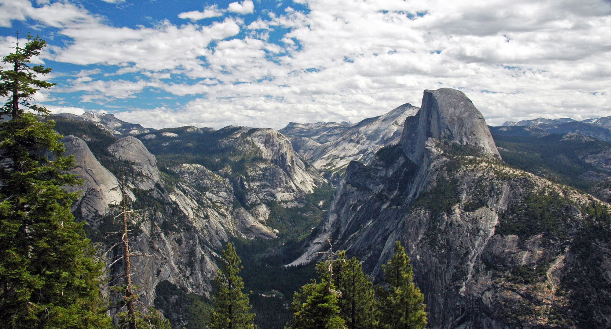 Photograph of Yosemite Valley taken from a high point in Yosemite National Park, California. The photo is taken looking down the valley with the peaks of the Sierra Nevada rising around it. Half Dome is in the center-right of the image.