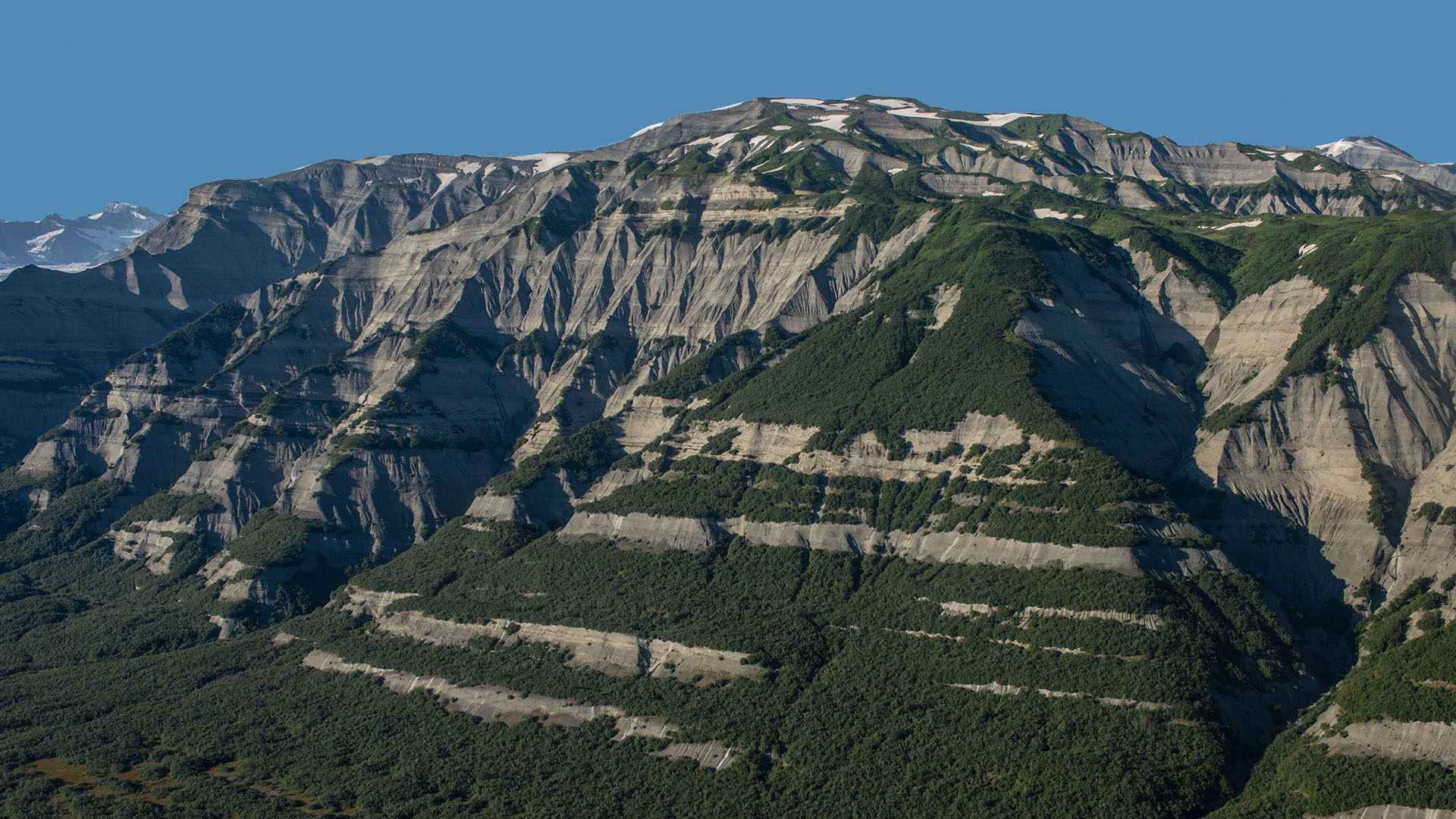Photograph of the Karr Hills, Yakutat Terrane, southern Alaska. The photo shows eroding hills partially of exposed brown rock and partially covered with green vegetation. Small patches of snow dot the tops of the hills.