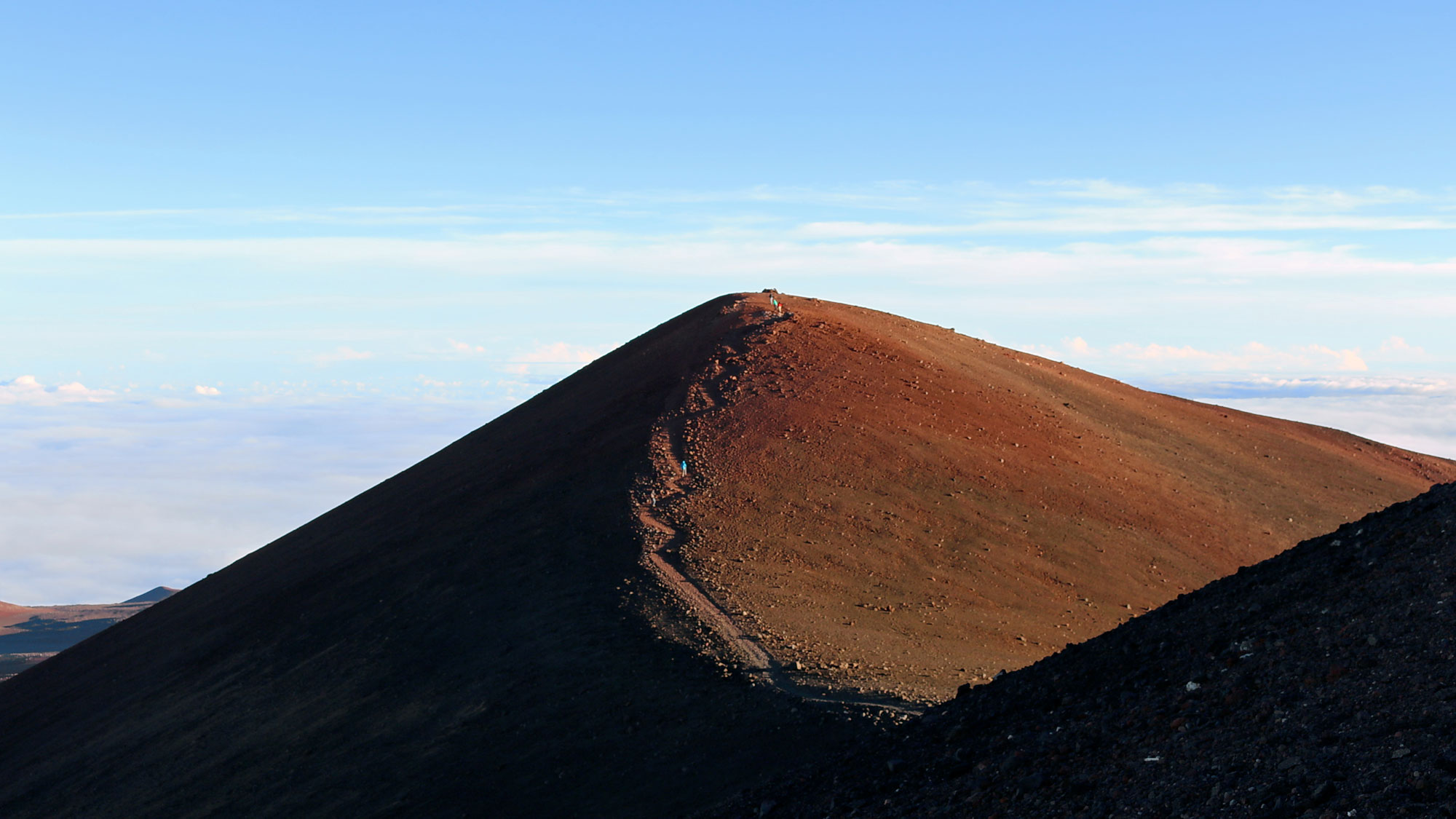 Photo of the summit trail on Mauna Kea. The photo shows a brown, barren, dry hill with a trail zigzagging up the nearest side.