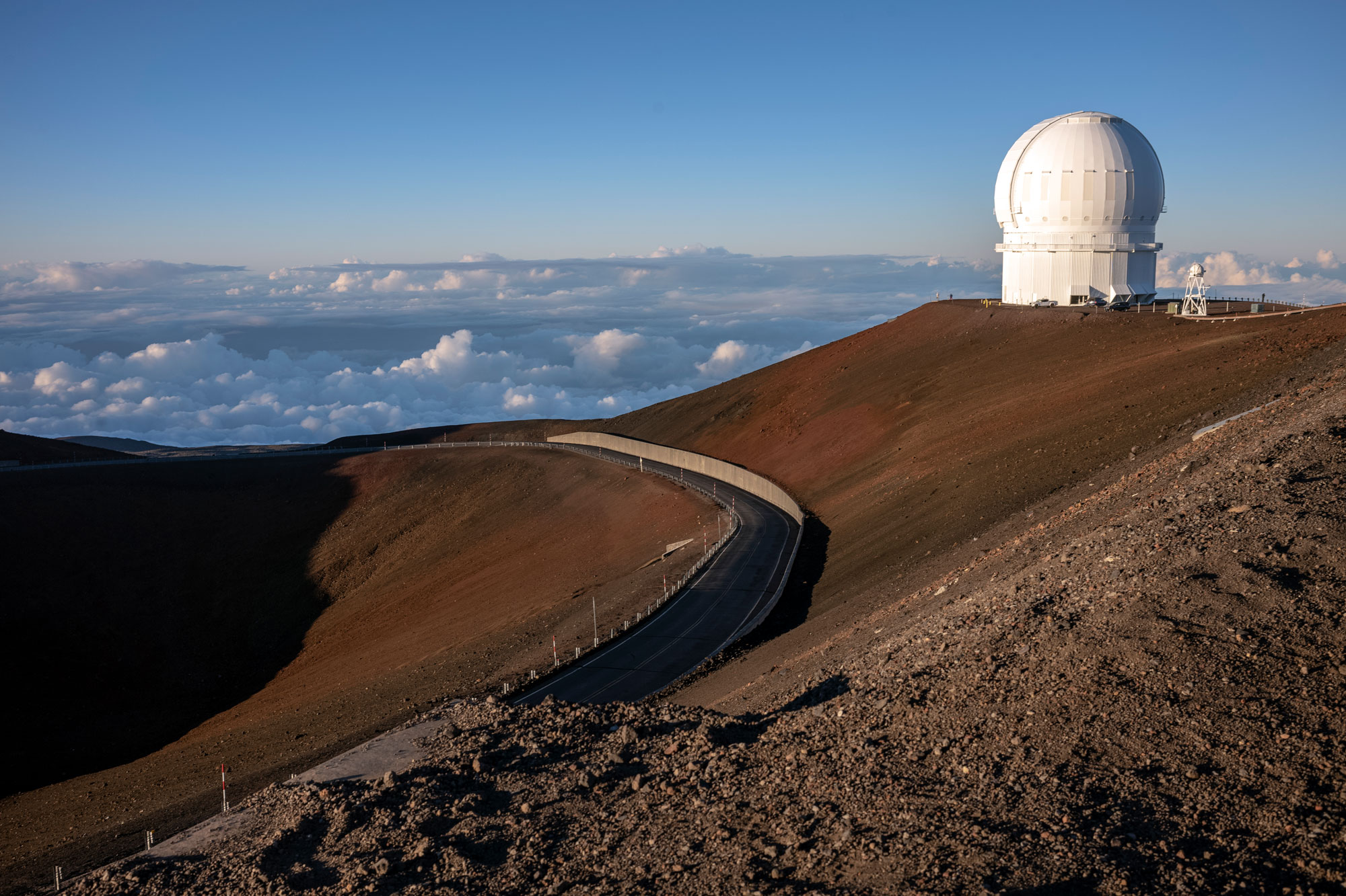 Photograph of the top of Mauna Loa showing the Mauna Loa Observatory. The top of the mountain is dry, brown, and barren of vegetation. The observatory is a cylindrical building topped with a white dome.