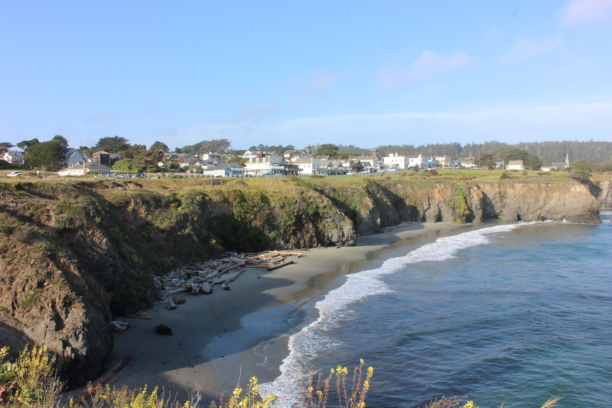 Photograph of Mendocino Village, California in 2015. The photo shows white buildings arranged on top of a low cliff along the ocean. In the foreground is a sandy beach at the base of the cliff.