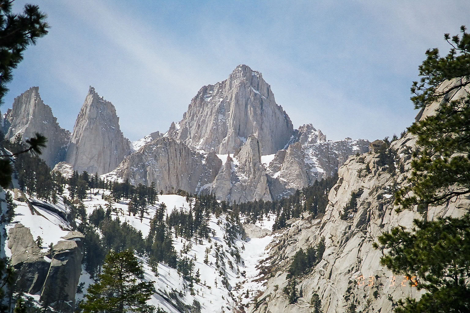 Photograph of the peak of Mount Whitney, the highest peak in California. The photo shows a triangular stone peak. In the foreground, a snowy slope is covered with conifers.